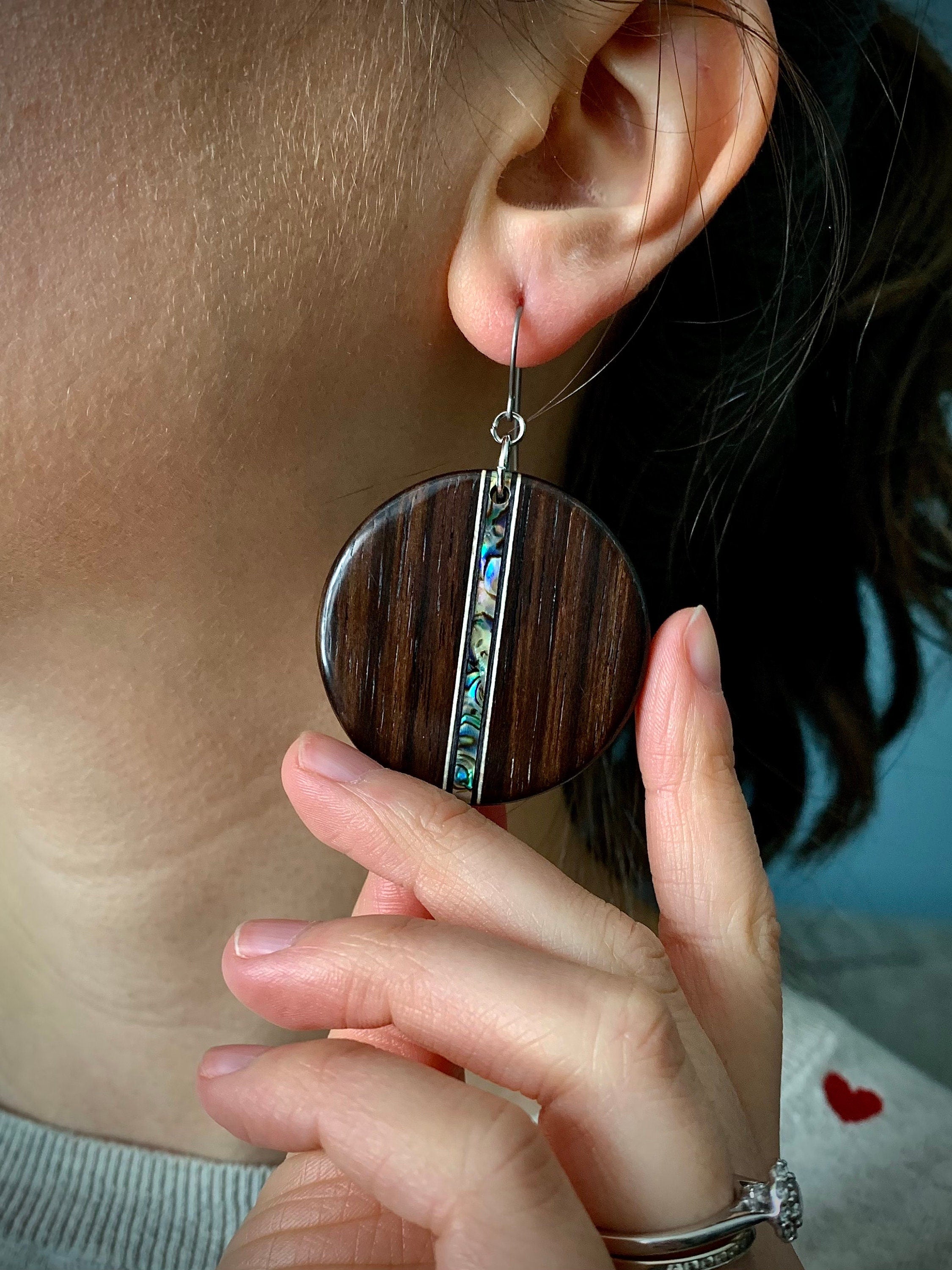 A round dangle earrings made of dark wood with a shimmering shell inlay running down the center on a model.