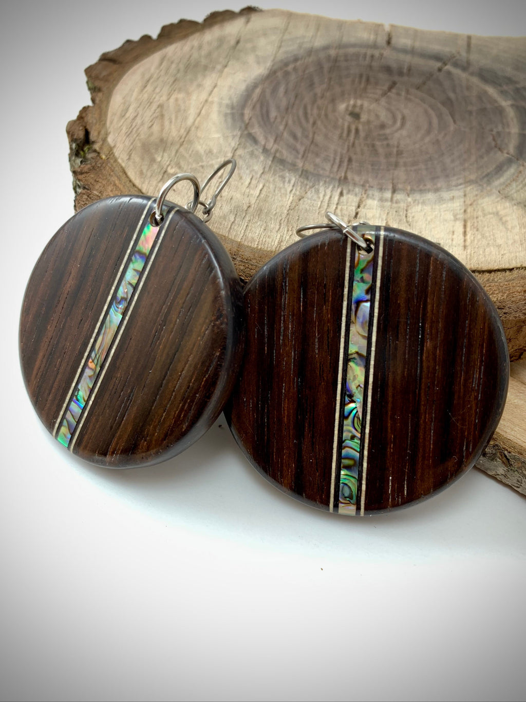 Two round earrings made of dark wood with a shimmering shell inlay running down the center leaning on a slab of wood.