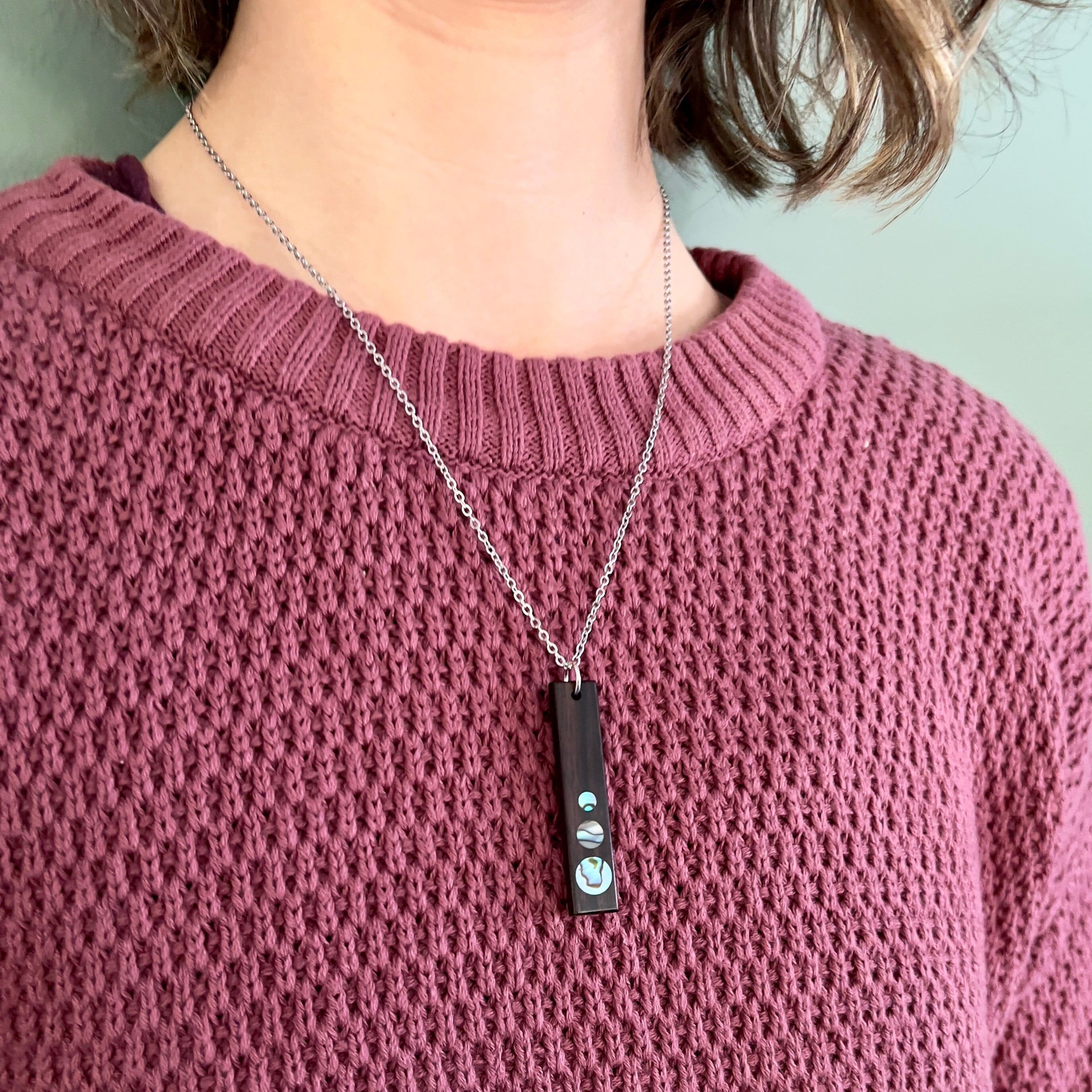 Close-up of a model wearing a knit sweater and a silver chain necklace with a rectangular pendant featuring three circular iridescent inlays.