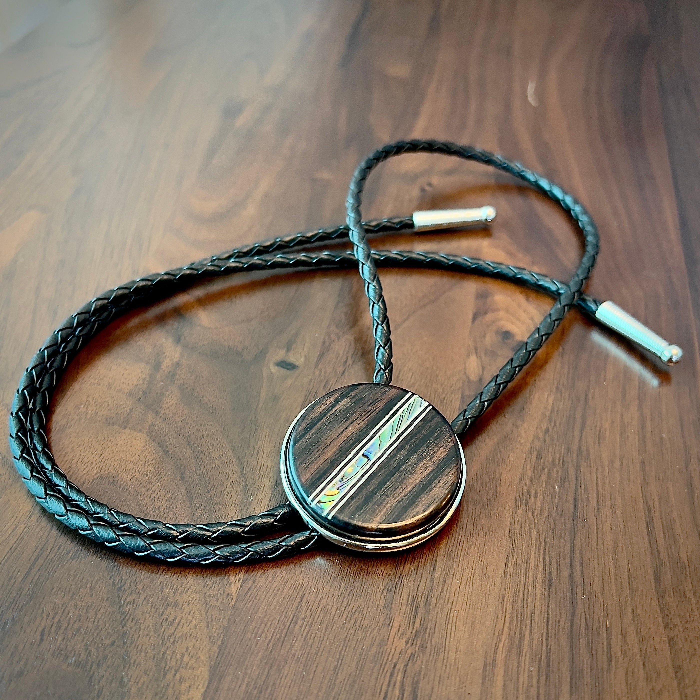 Bolo tie with a round wooden slide with iridescent vertical inlay strip and black braided cords on a wooden surface.