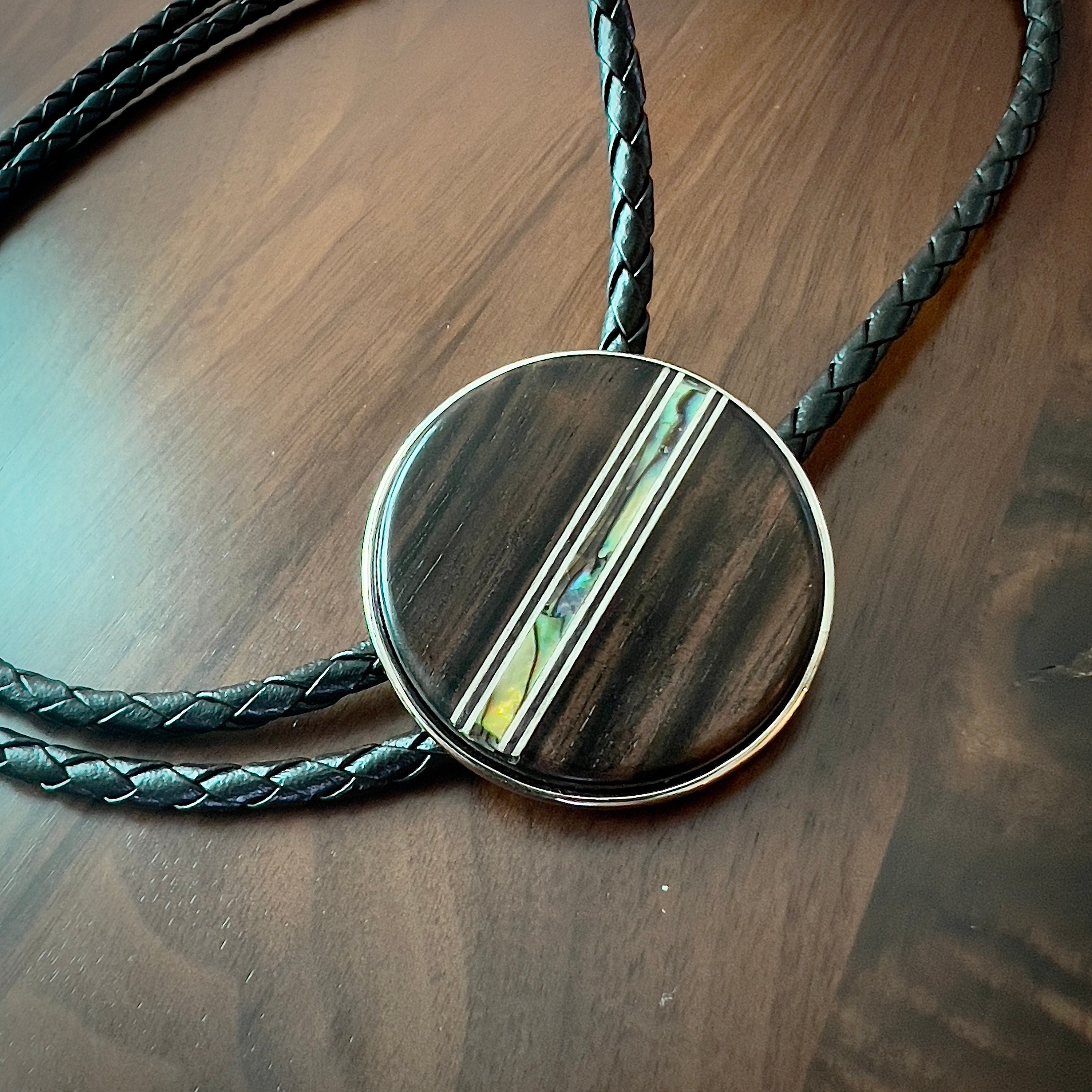 Circular bolo slide with a dark wooden texture and an iridescent inlay, on a black braided cord.