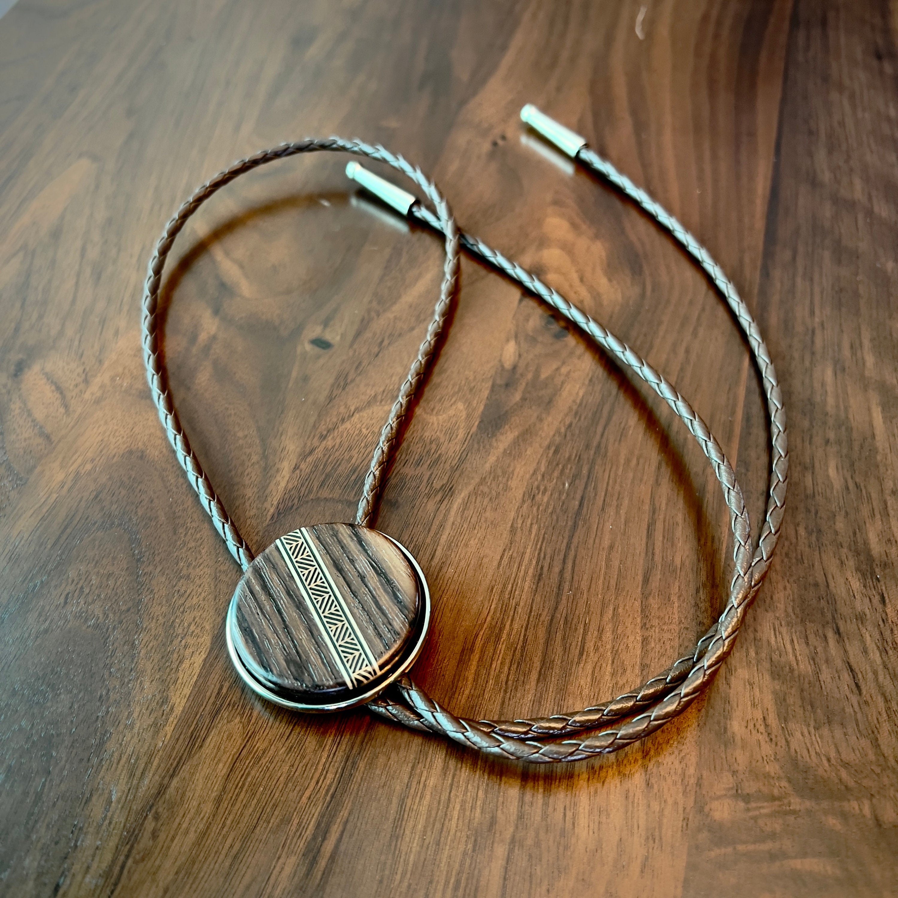 A bolo tie with a dark wood circular centerpiece featuring a tan-toned geometric inlay. The braided brown leather cords complement the wood’s natural texture, resting on a matching wooden surface to highlight the tie’s handcrafted design.