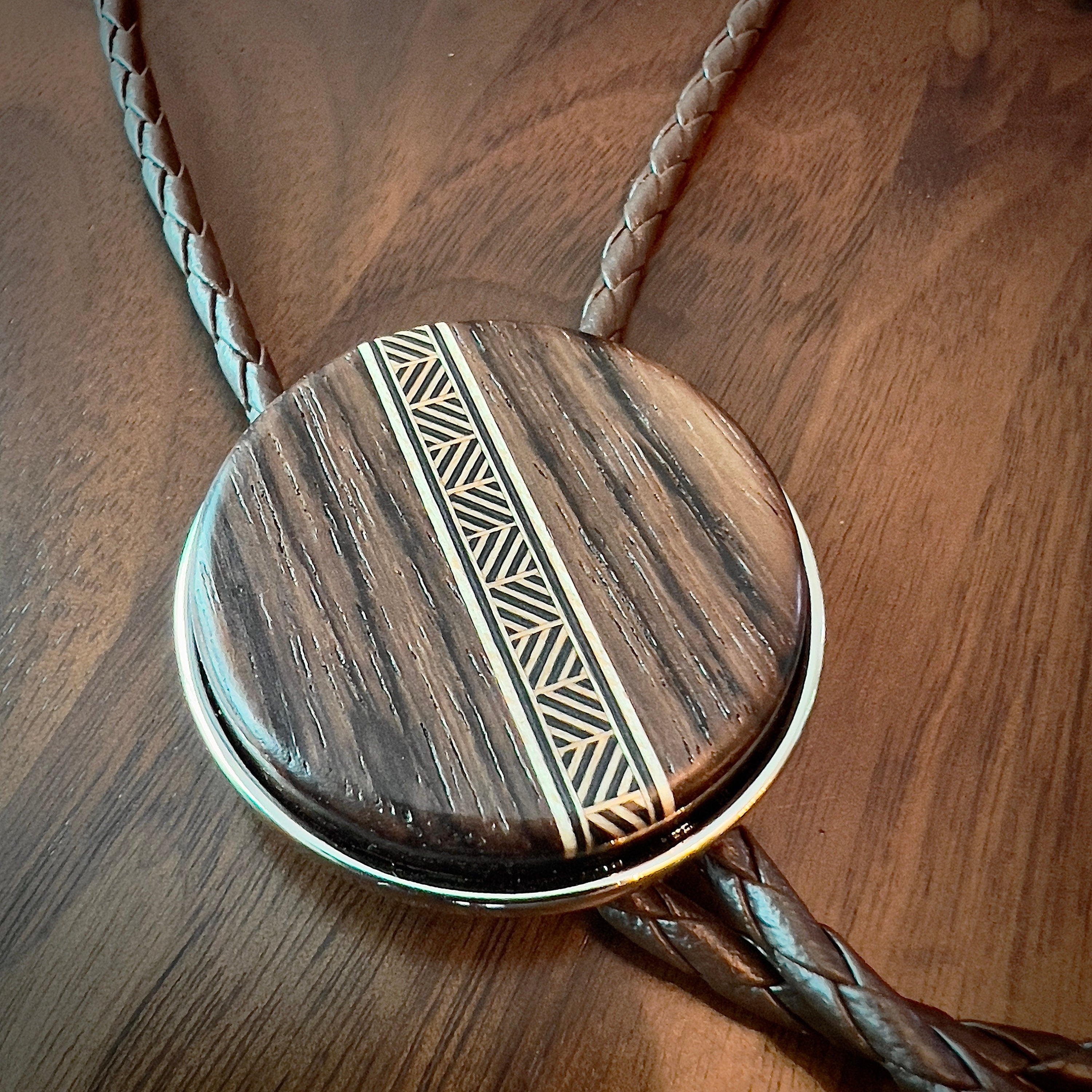 A bolo tie with a dark wood circular centerpiece featuring a tan-toned geometric inlay. The braided brown leather cords complement the wood’s natural texture, resting on a matching wooden surface to highlight the tie’s handcrafted design.