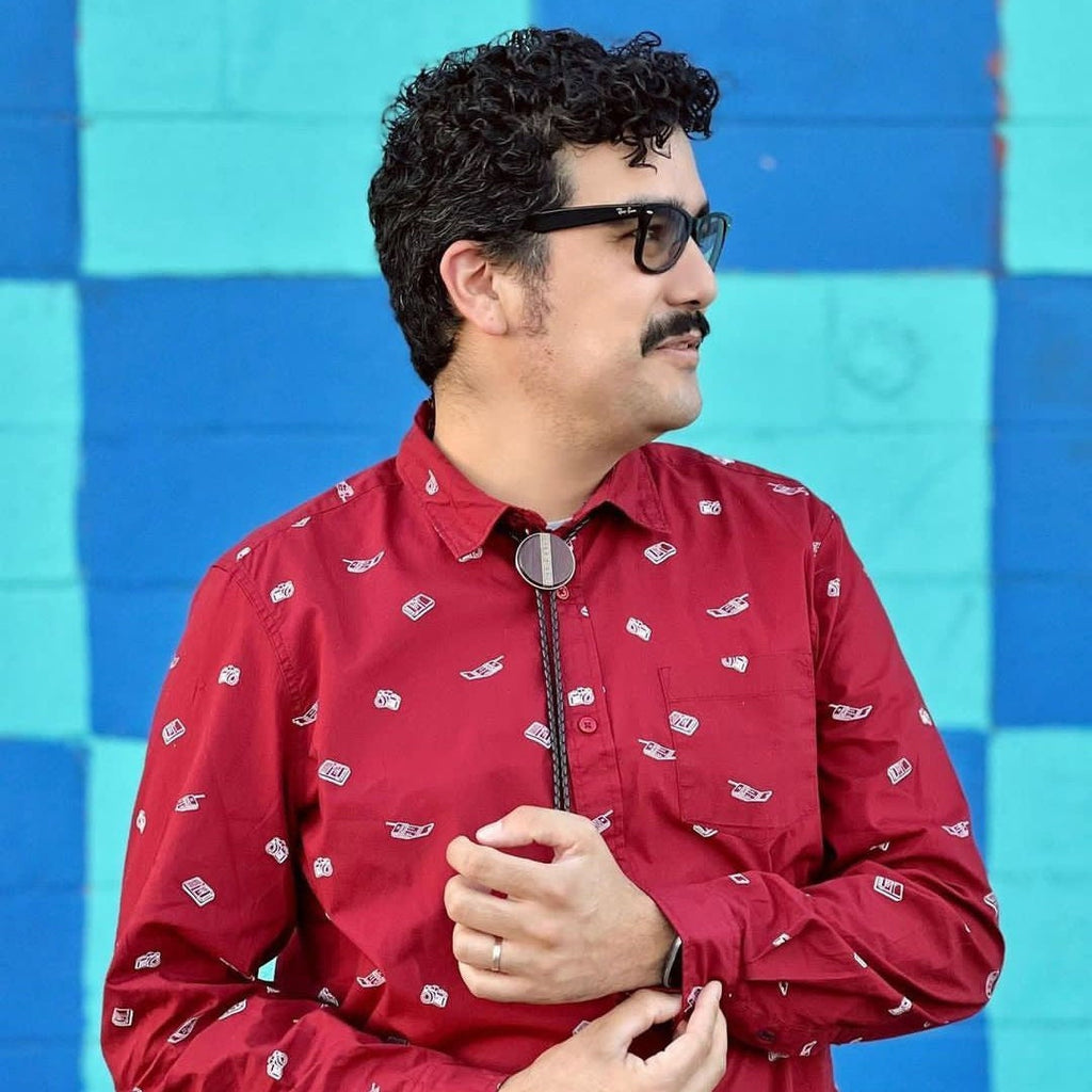 A man wearing a bolo tie with a dark wood circular centerpiece and braided black leather cords. He pairs it with a red button-up shirt and black glasses. The background is a bold, colorful blue checkered wall, adding a vibrant contrast.