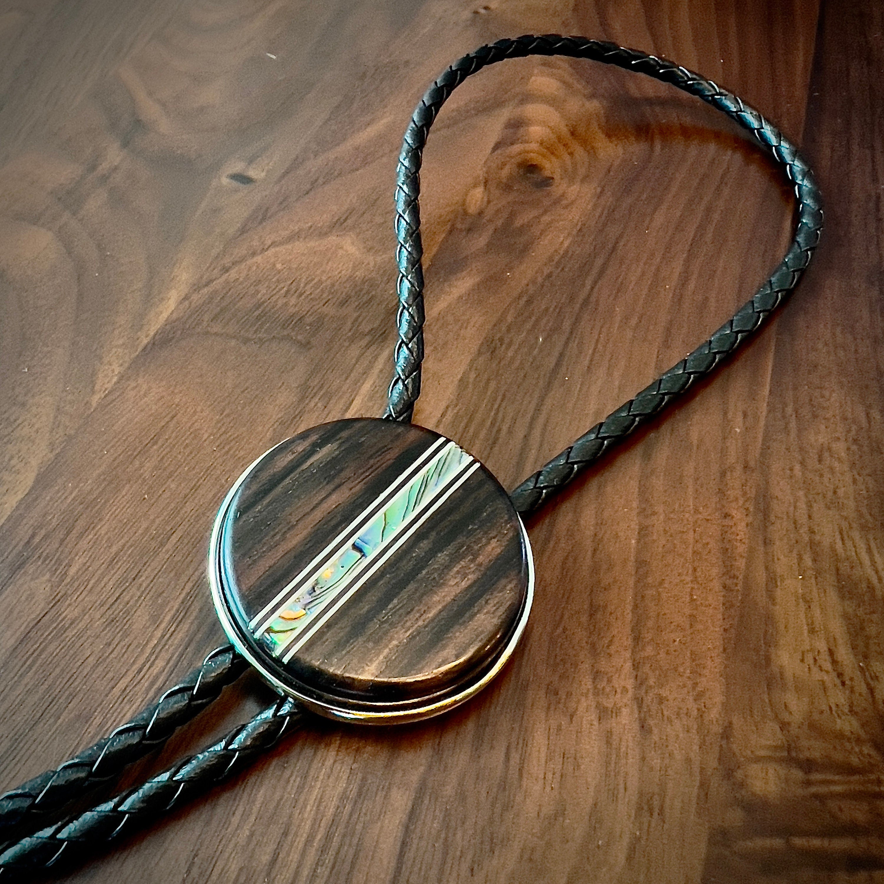 Circular bolo slide with a dark wooden texture and an iridescent inlay, on a black braided cord.