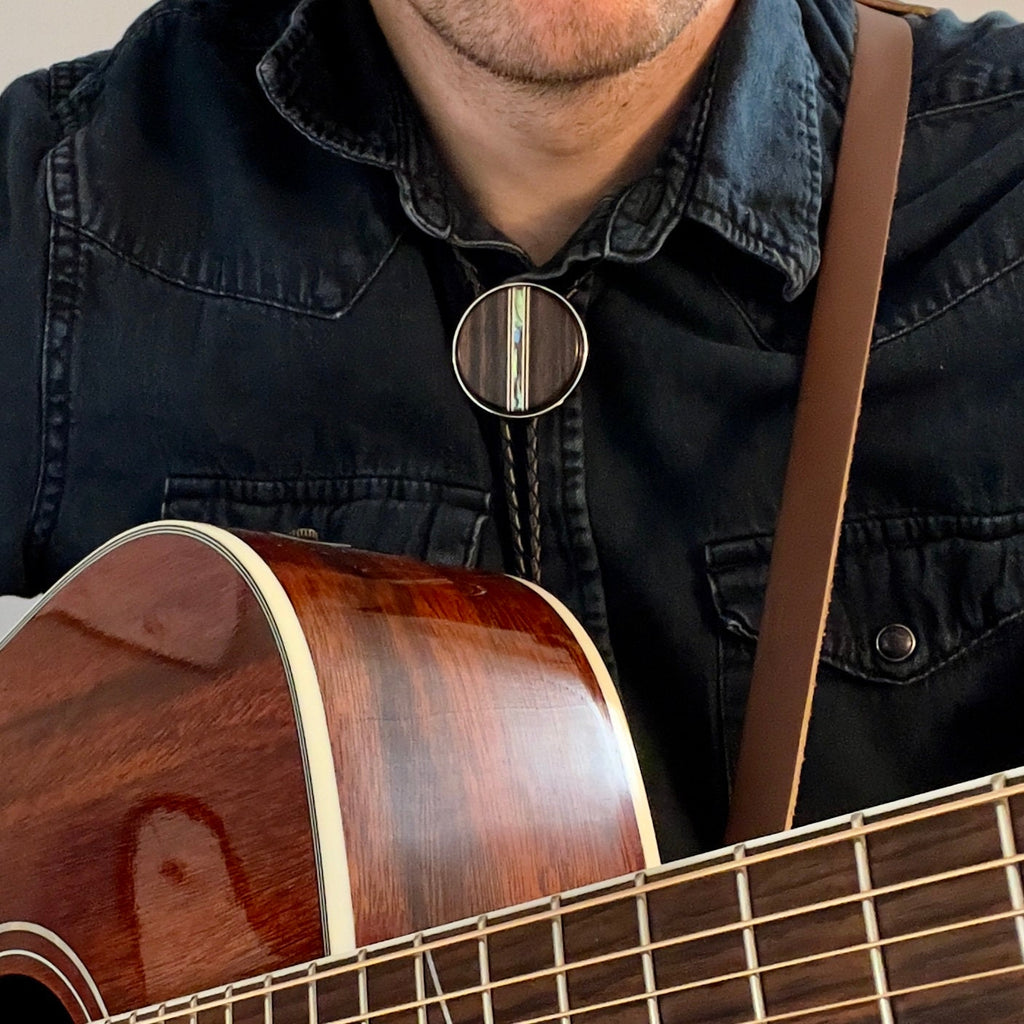 Person wearing a dark denim shirt and bolo tie, holding a wooden acoustic guitar.