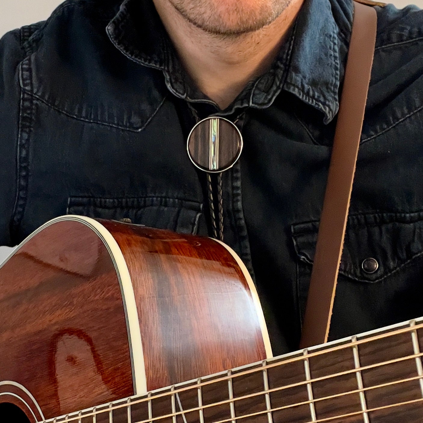 Person wearing a dark denim shirt and bolo tie, holding a wooden acoustic guitar.