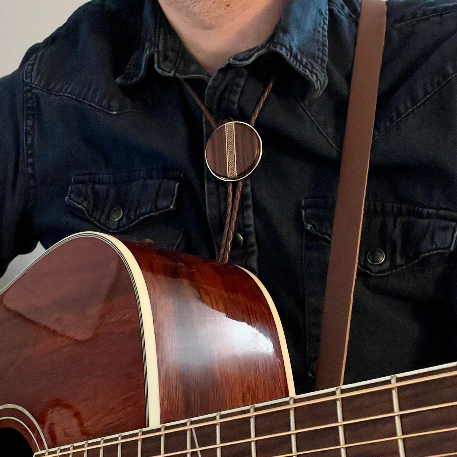 A person wearing a bolo tie with a wood and centerpiece featuring intricate geometric inlay. They are dressed in a dark denim shirt, adding a casual, rustic style.