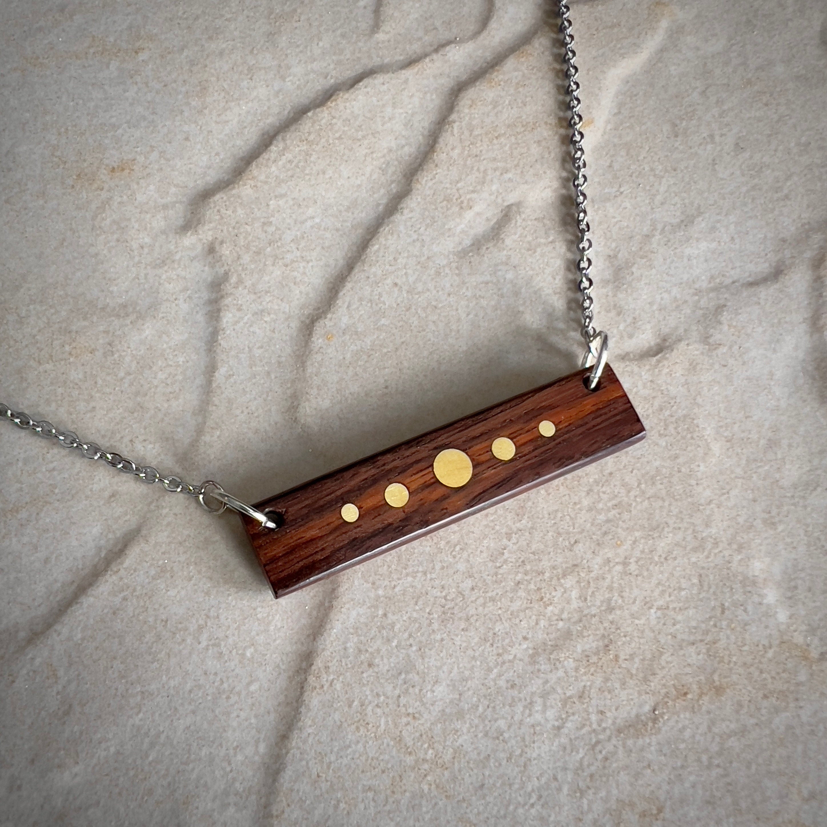 A rectangular wooden pendant with brass inlay dots of varying sizes hangs from a silver chain against a stone surface. The design resembles a minimalist planetary alignment. The wood has a rich, dark tone, contrasting with the bright brass details.