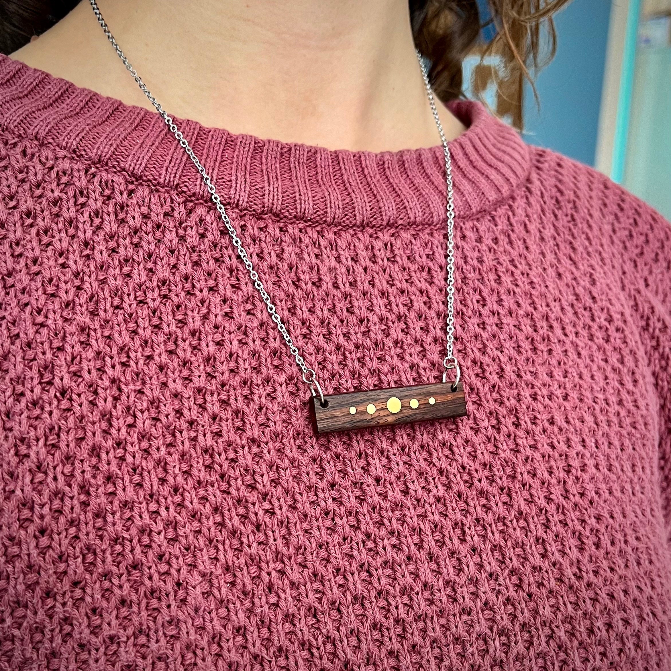 A person wears a silver-chained necklace with a rectangular wooden pendant featuring brass inlay dots. The pendant rests against a pink sweater. The design resembles a planetary alignment, with the dark wood contrasting against the brass details.