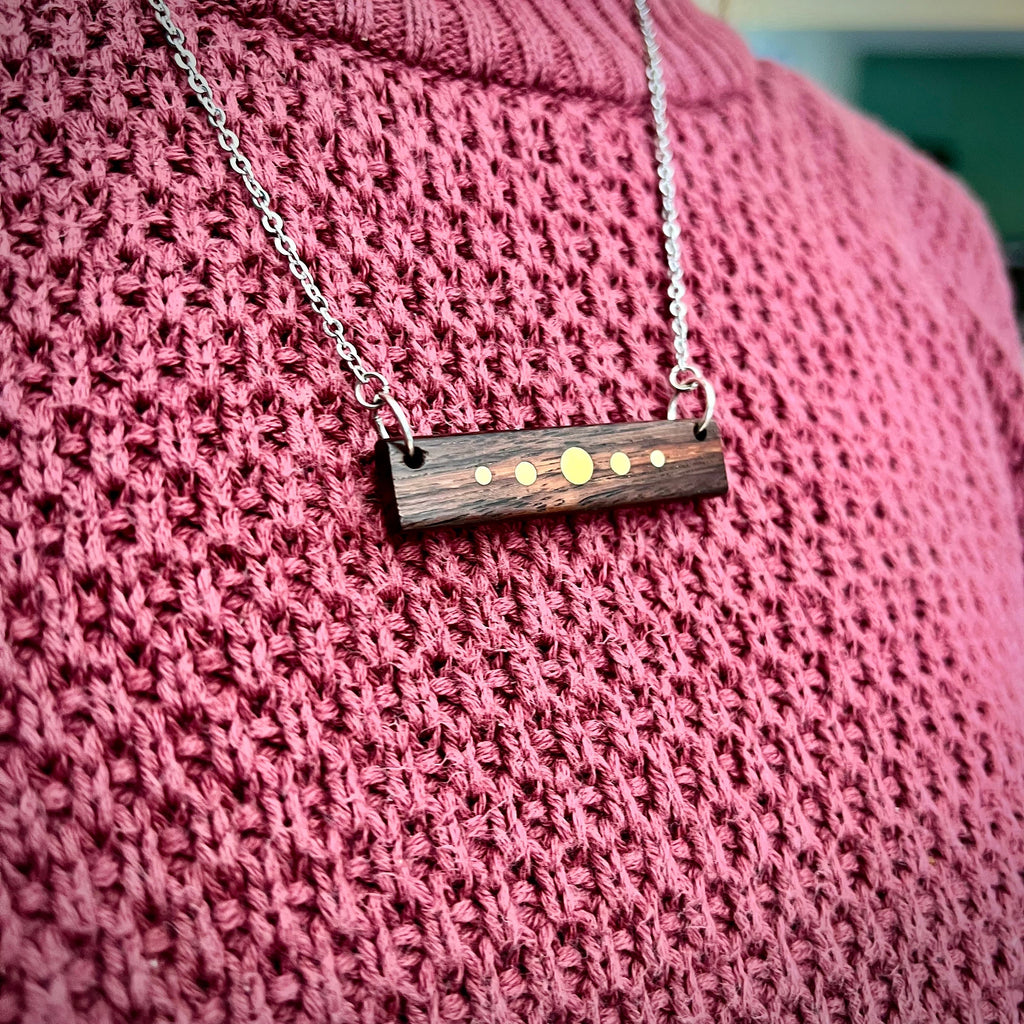 A person wears a silver-chained necklace with a rectangular wooden pendant featuring brass inlay dots. The pendant rests against a pink sweater. The design resembles a planetary alignment, with the dark wood contrasting against the brass details.
