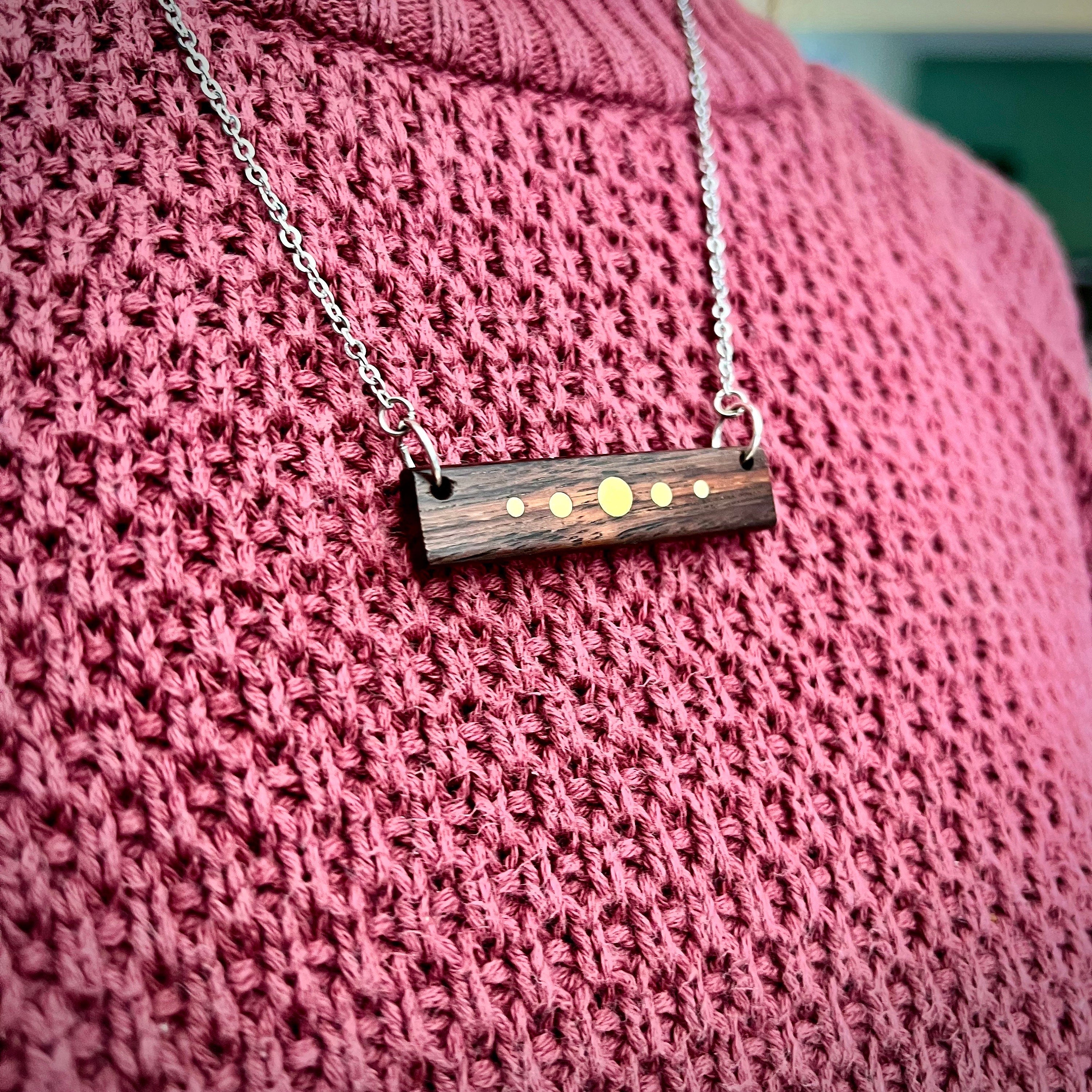 A person wears a silver-chained necklace with a rectangular wooden pendant featuring brass inlay dots. The pendant rests against a pink sweater. The design resembles a planetary alignment, with the dark wood contrasting against the brass details.