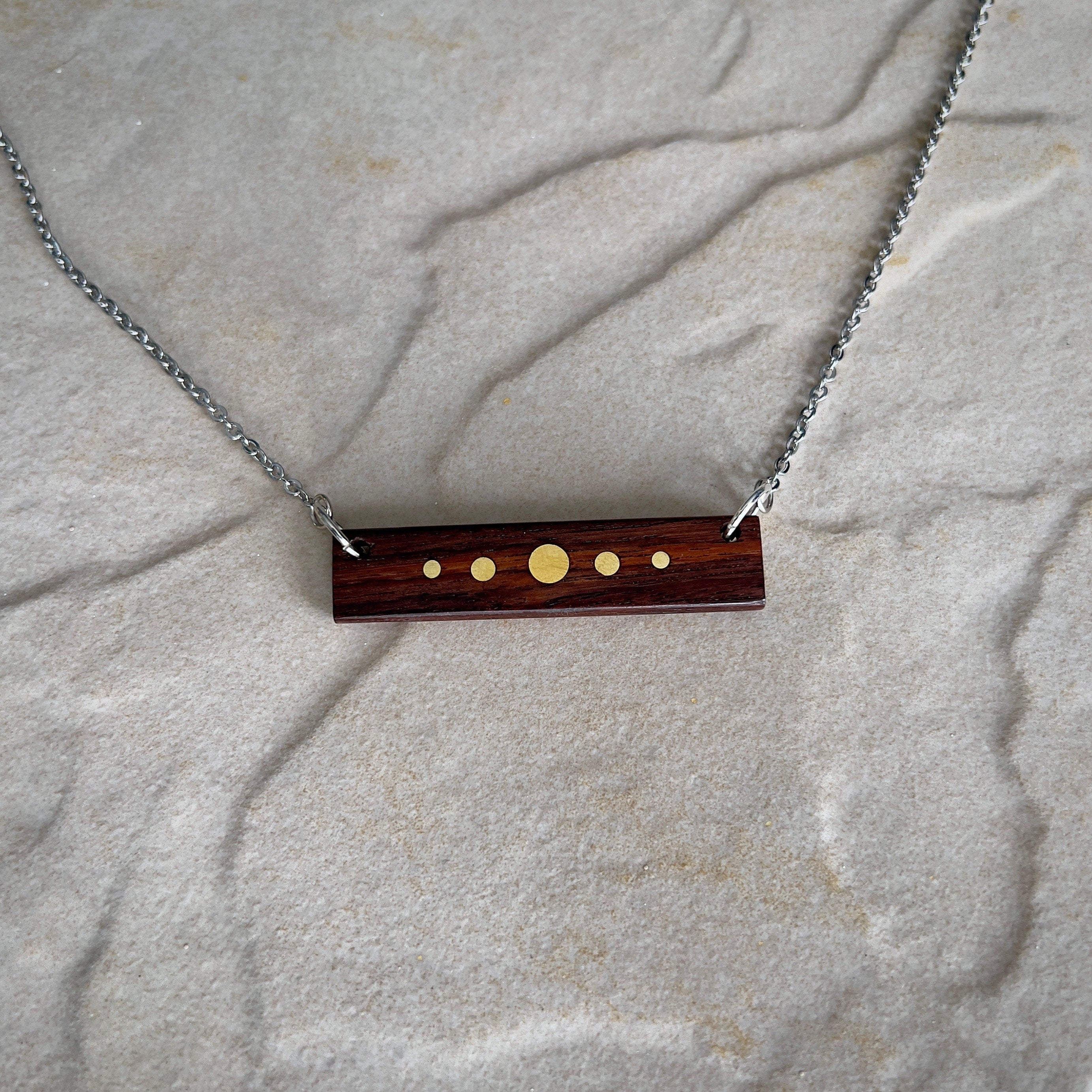 A rectangular wooden pendant with brass inlay dots of varying sizes hangs from a silver chain against a stone surface. The design resembles a minimalist planetary alignment. The wood has a rich, dark tone, contrasting with the bright brass details.