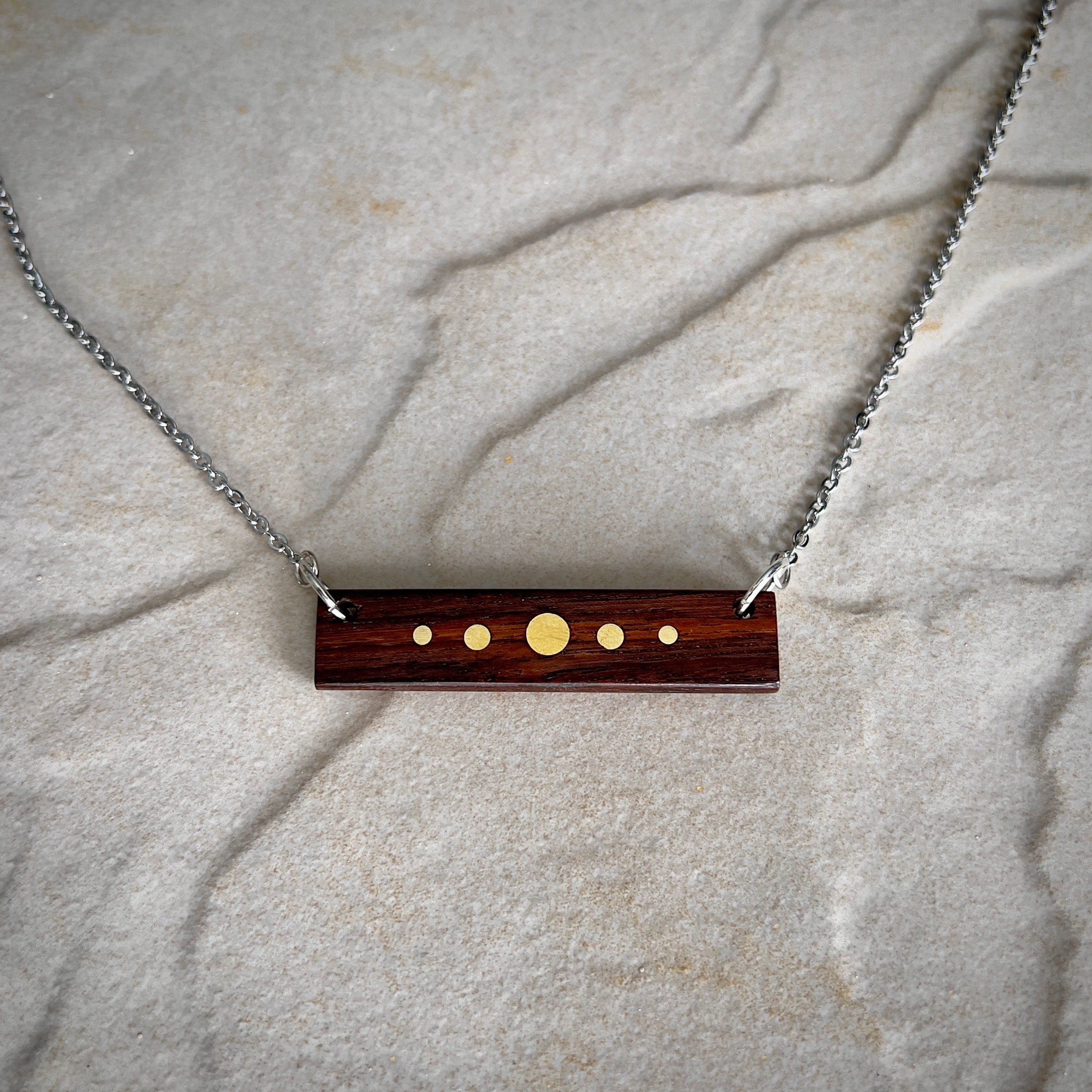 A rectangular wooden pendant with brass inlay dots of varying sizes hangs from a silver chain against a stone surface. The design resembles a minimalist planetary alignment. The wood has a rich, dark tone, contrasting with the bright brass details.