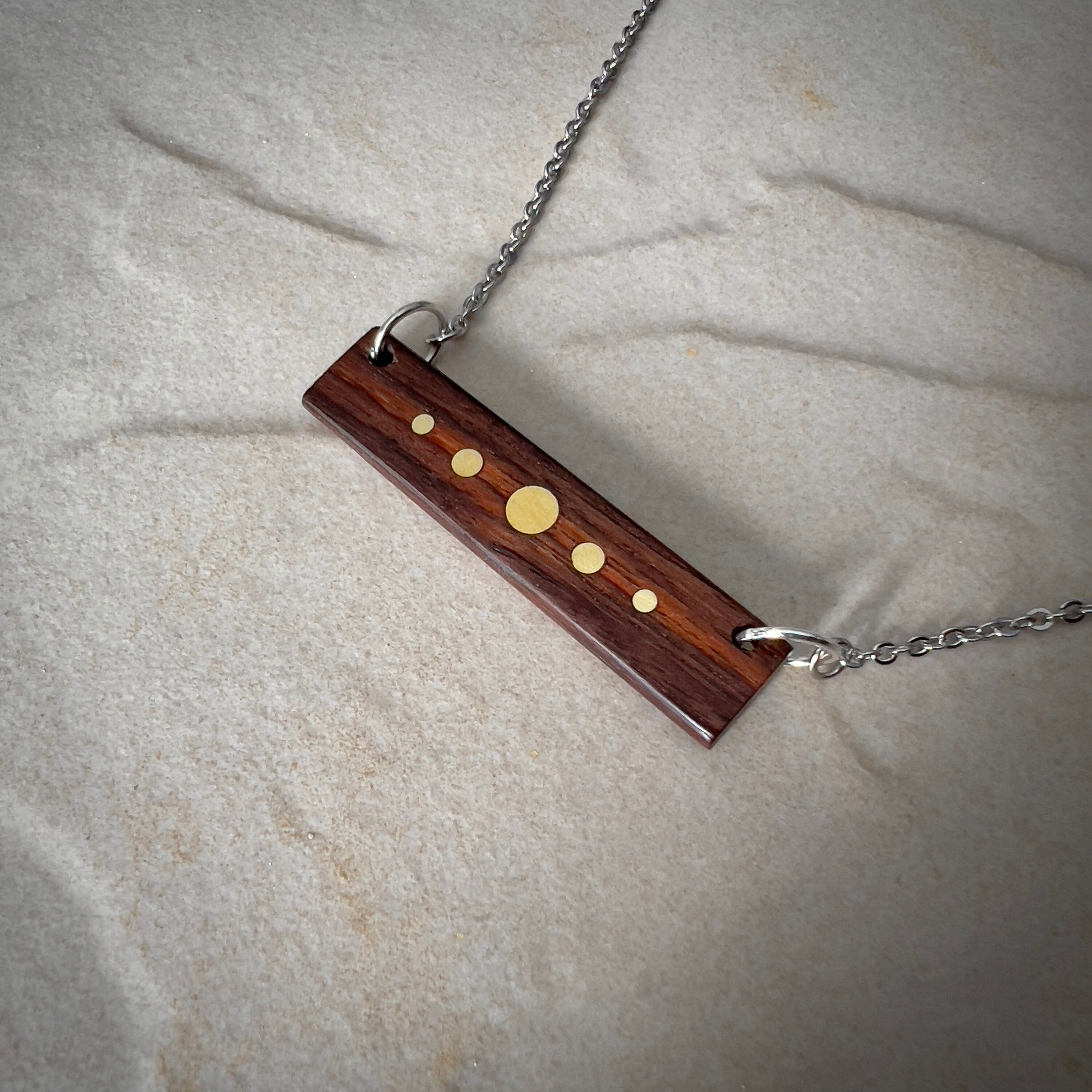 A rectangular wooden pendant with brass inlay dots of varying sizes hangs from a silver chain against a stone surface. The design resembles a minimalist planetary alignment. The wood has a rich, dark tone, contrasting with the bright brass details.