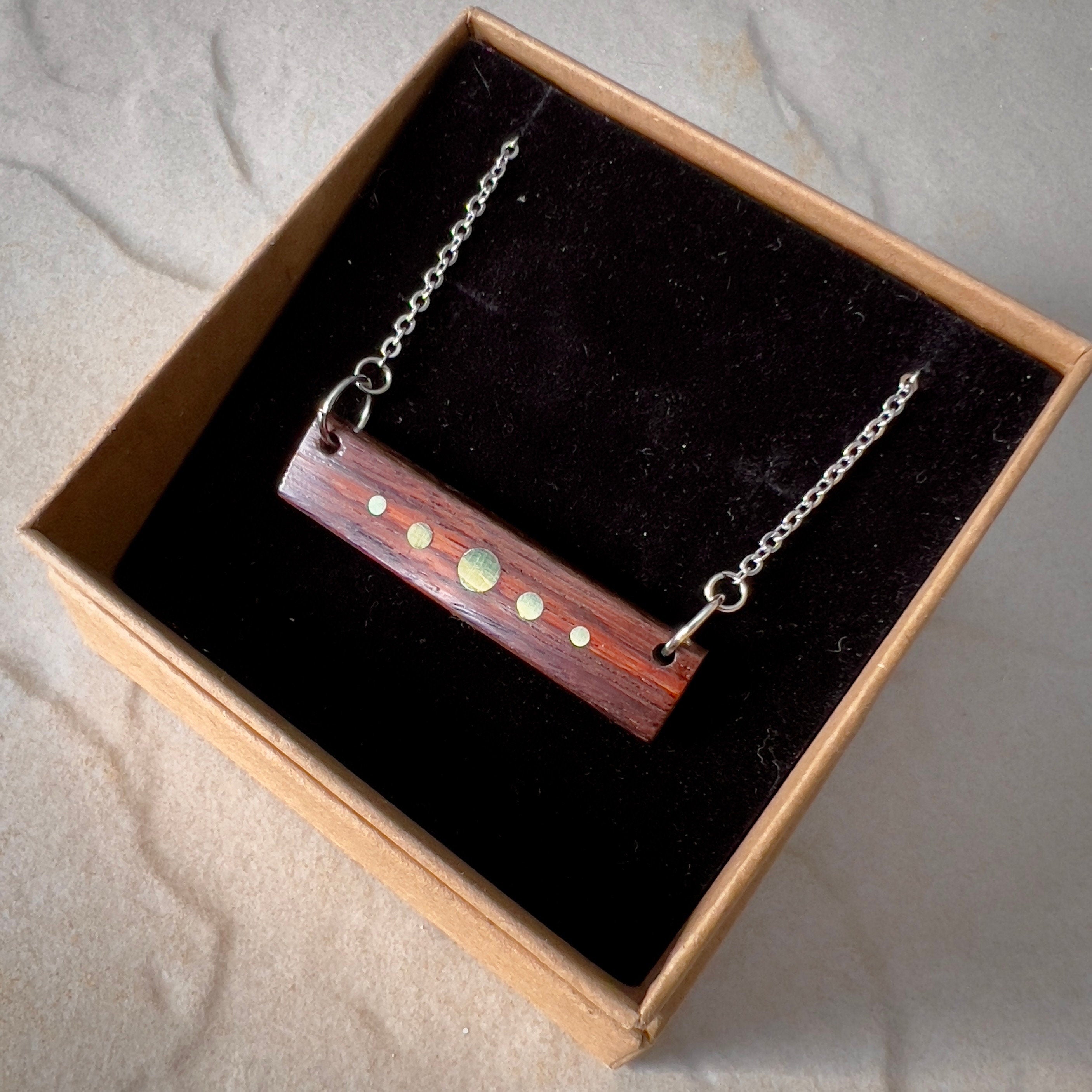 A rectangular wooden pendant with brass inlay dots hangs from a silver chain, displayed in a black velvet-lined cardboard box. The dark wood contrasts with the bright brass details, resembling a planetary design. The box rests on a stone surface.