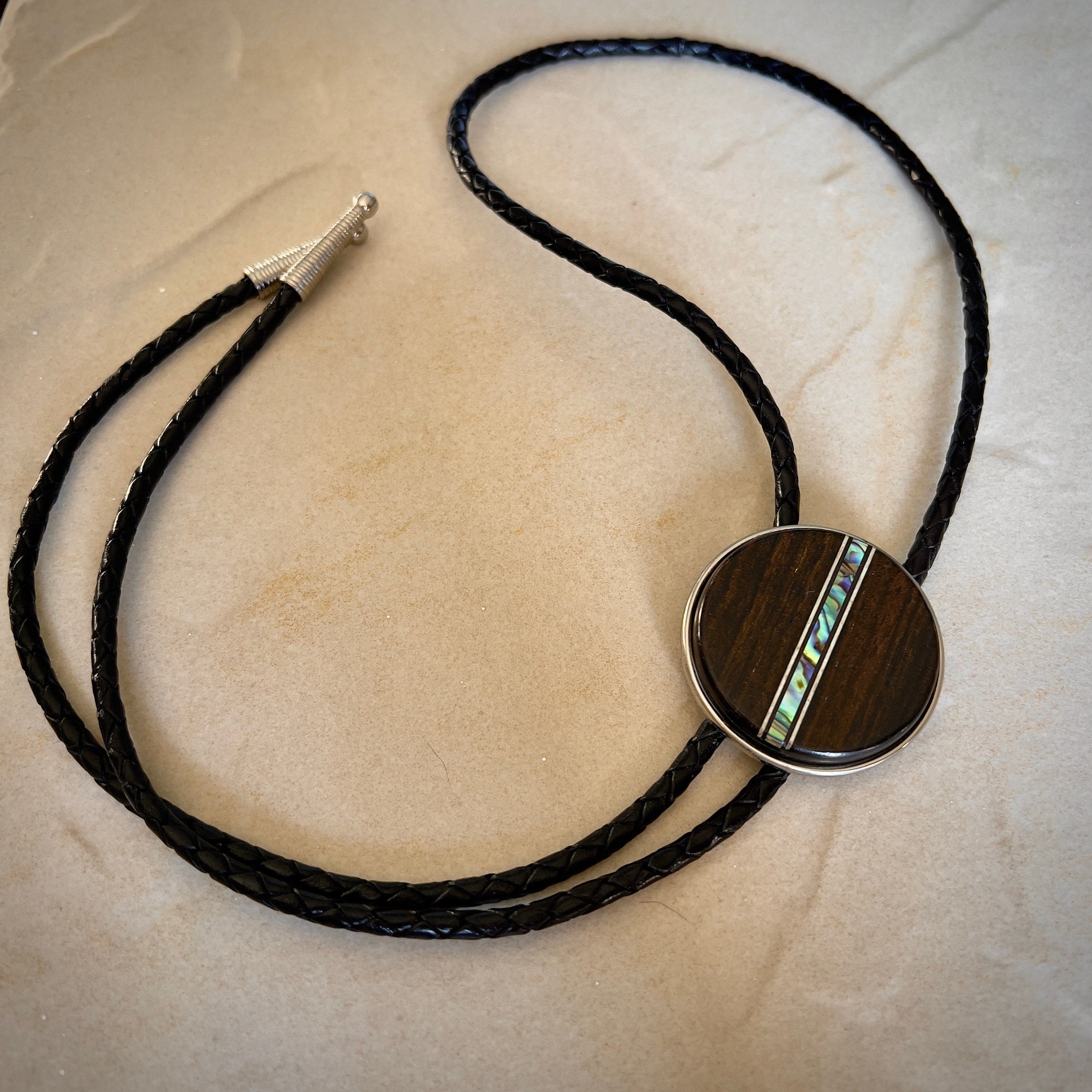 Bolo tie with a round wooden slide with iridescent vertical inlay strip and black braided cords on a stone surface.
