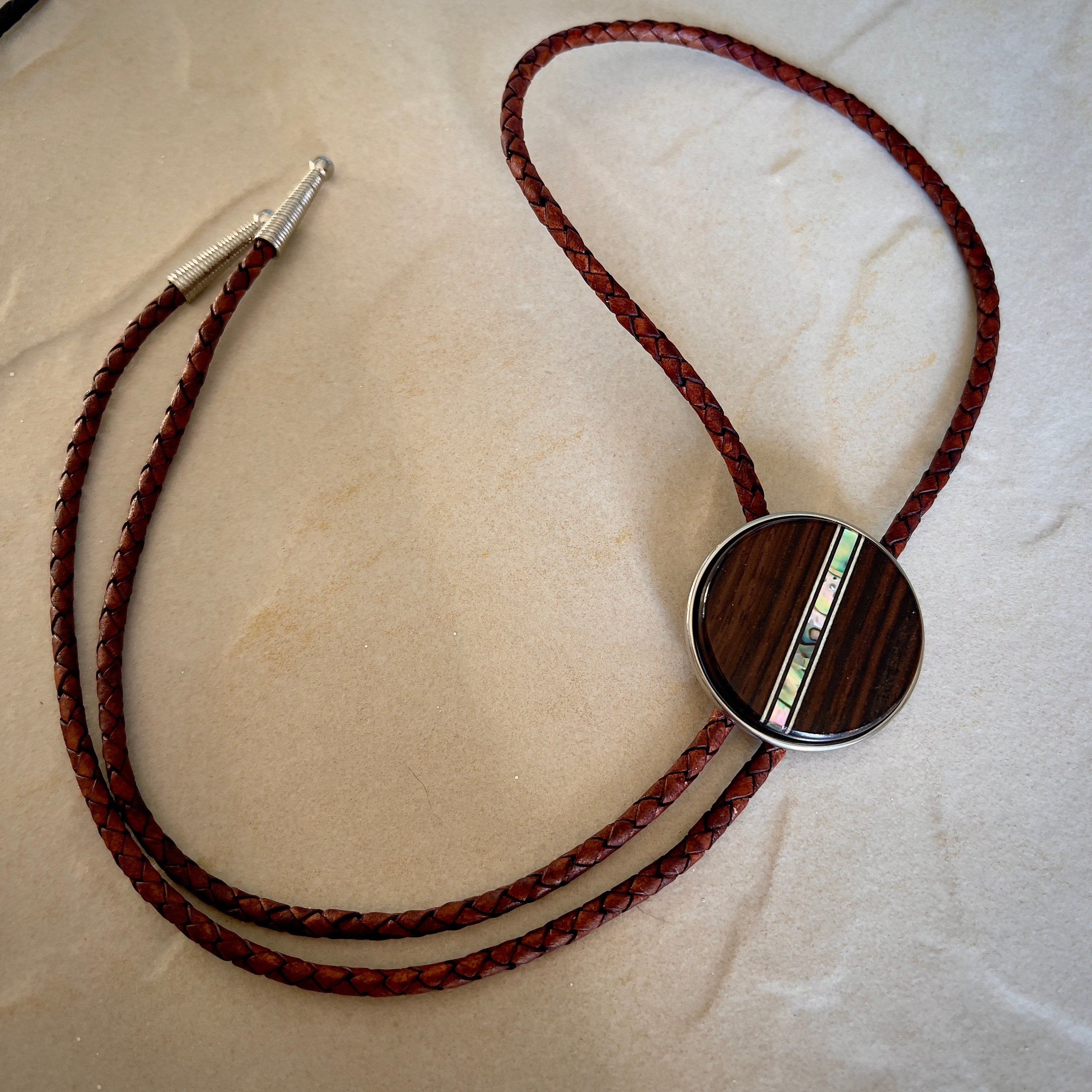 Bolo tie with a round wooden slide with iridescent vertical inlay strip and brown braided cords on a stone surface.