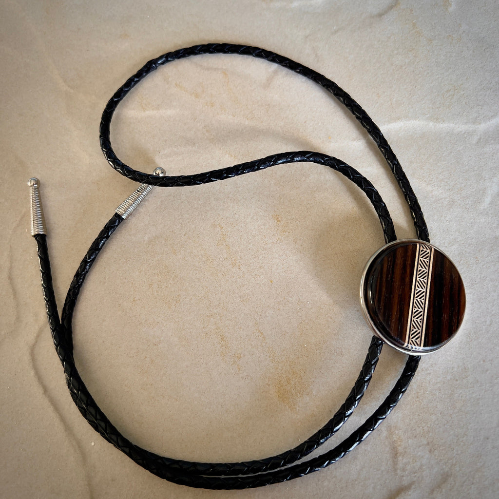 A bolo tie with a dark wood circular centerpiece featuring a tan-toned geometric inlay. The braided brown leather cords complement the wood’s natural texture, resting on a stone surface to highlight the tie’s handcrafted design.