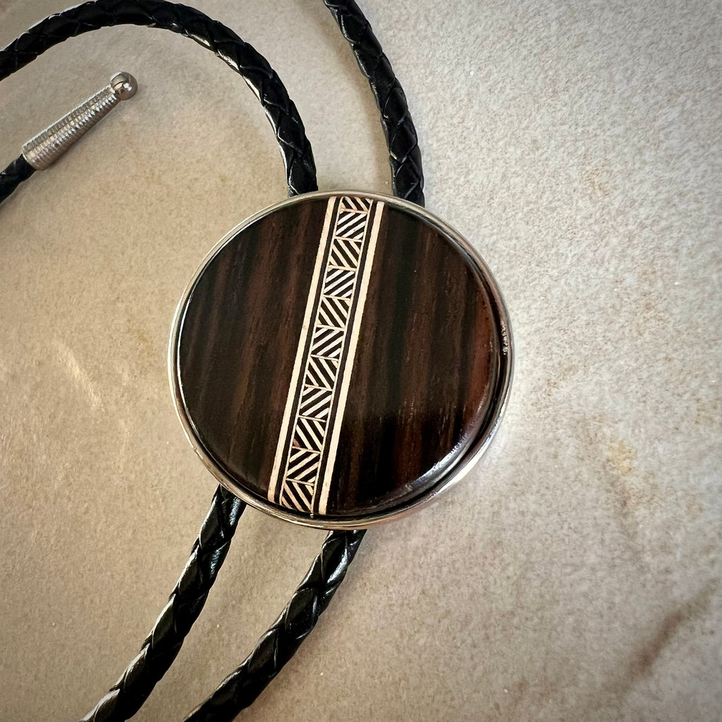 A bolo tie with a dark wood circular centerpiece featuring a tan-toned geometric inlay. The braided brown leather cords complement the wood’s natural texture, resting on a stone surface to highlight the tie’s handcrafted design.