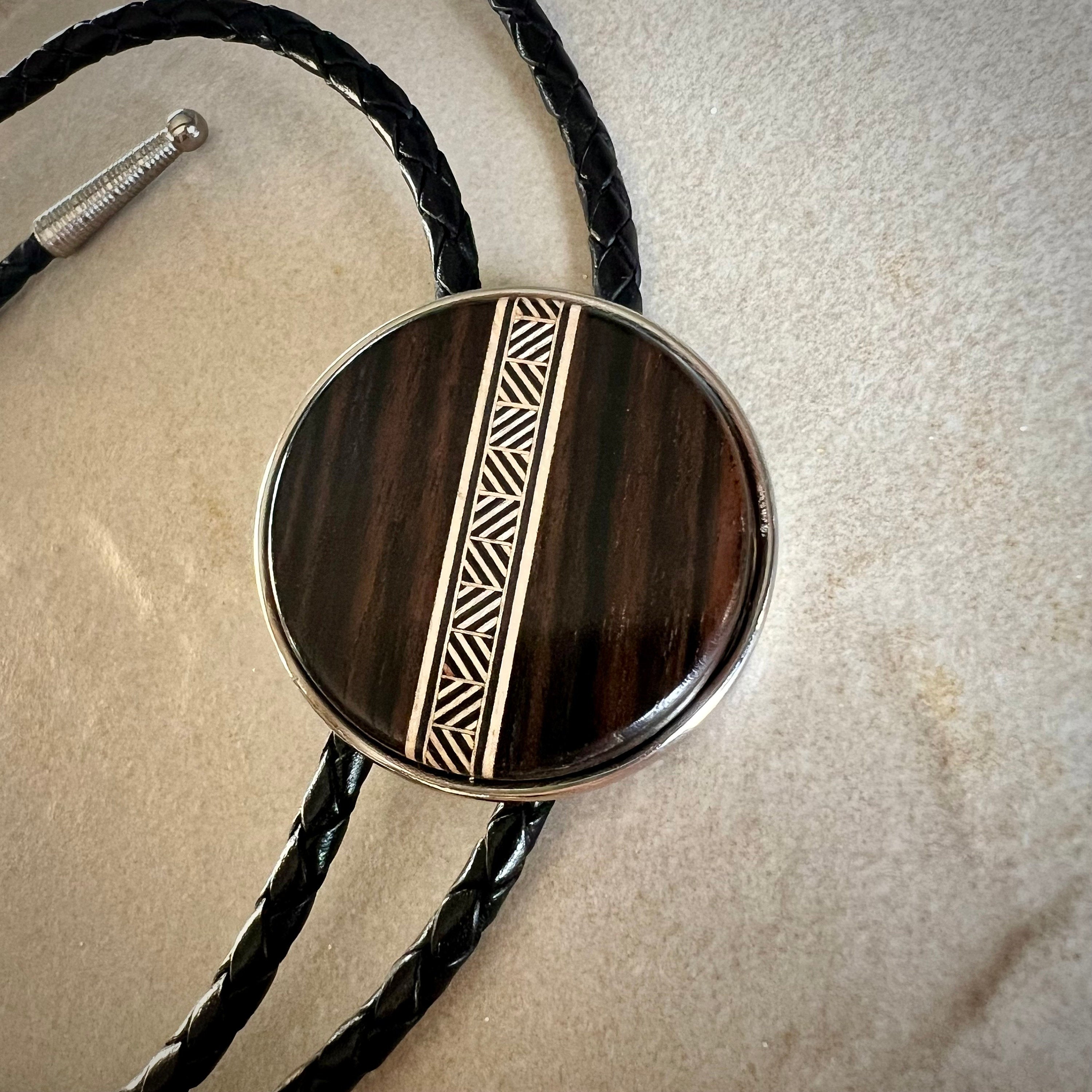 A bolo tie with a dark wood circular centerpiece featuring a tan-toned geometric inlay. The braided brown leather cords complement the wood’s natural texture, resting on a stone surface to highlight the tie’s handcrafted design.