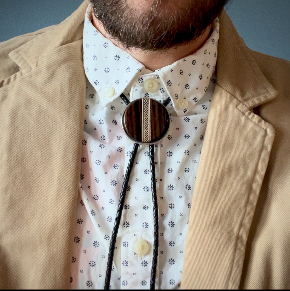 A bolo tie with a dark wood circular centerpiece featuring a tan-toned geometric inlay. It is worn with a patterned white shirt and beige jacket, with braided black leather cords completing the accessory’s refined, Western-inspired style.
