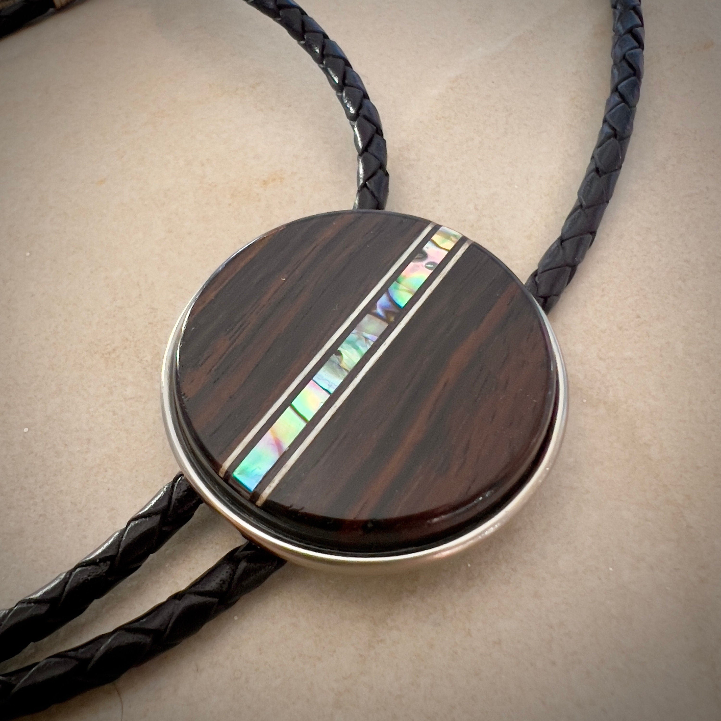 Circular bolo slide with a dark wooden texture and an iridescent inlay, on a black braided cord.