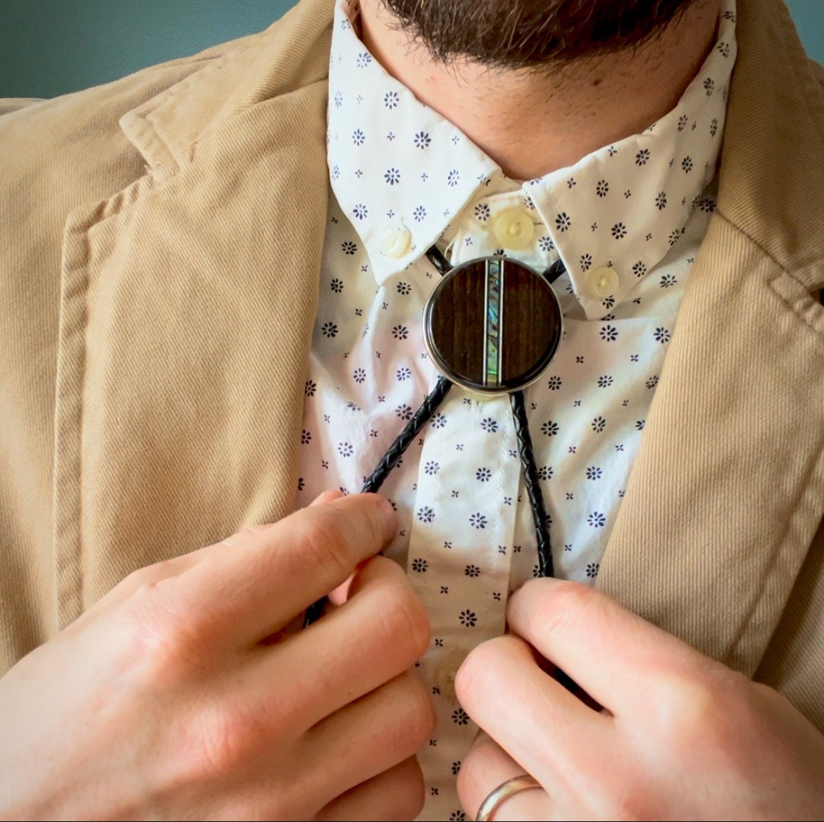 Close-up of a person adjusting a bolo tie over a patterned white shirt and tan blazer.