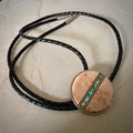 Bolo tie with a round wooden slide with iridescent vertical inlay strip and black braided cords on a wooden surface.
