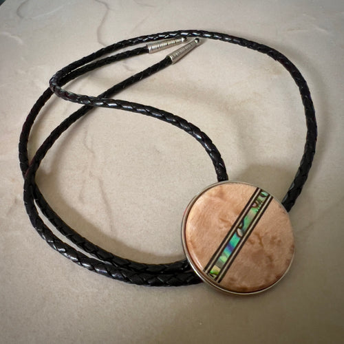 Bolo tie with a round wooden slide with iridescent vertical inlay strip and black braided cords on a wooden surface.