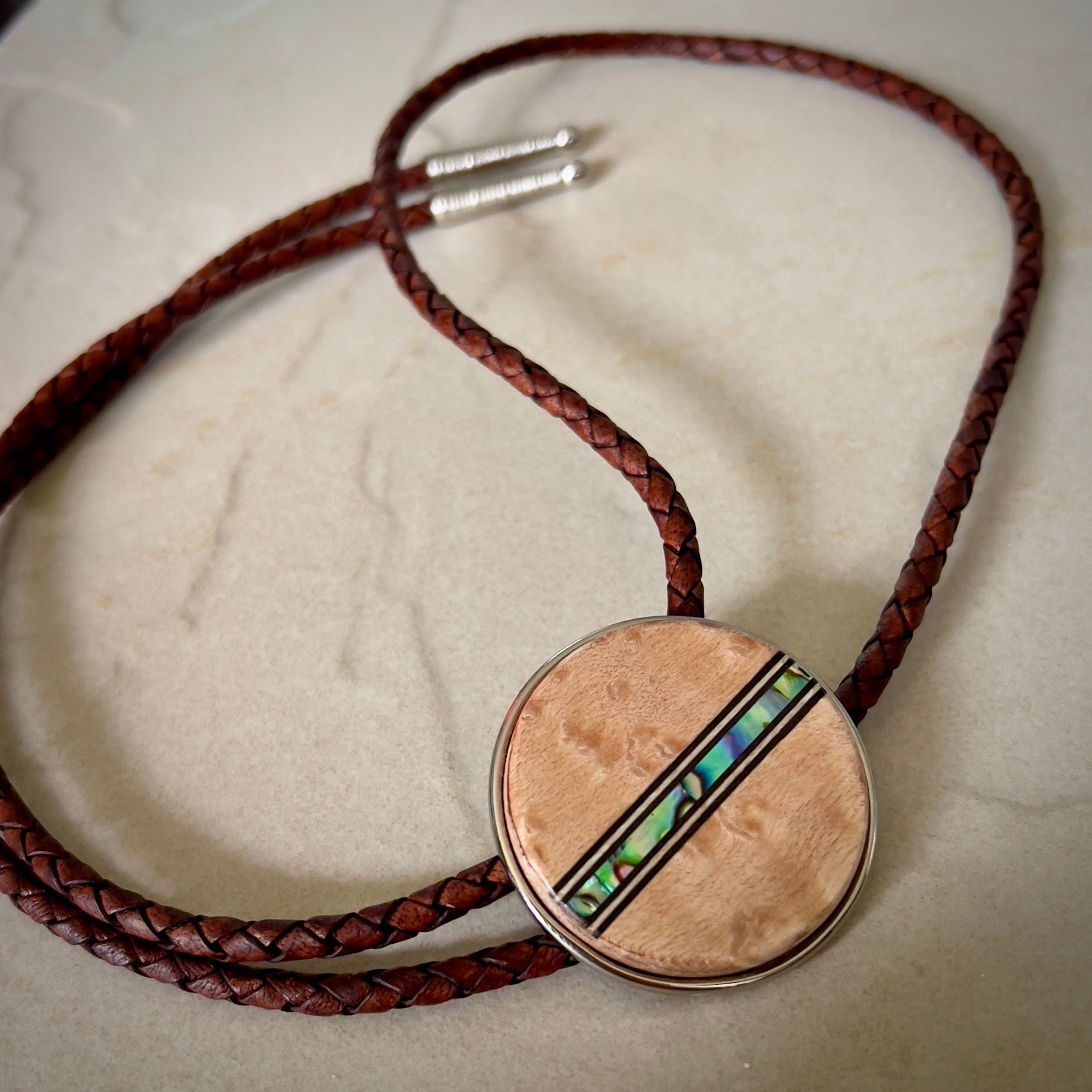 Bolo tie with a round wooden slide with iridescent vertical inlay strip and brown braided cords on a wooden surface.