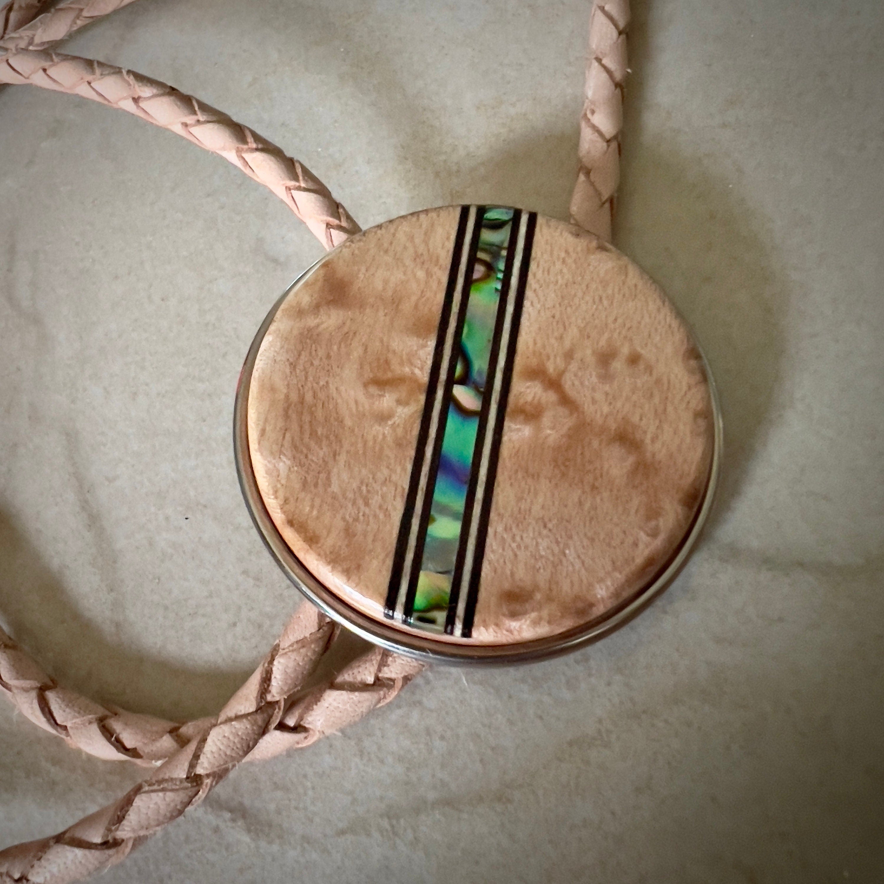 Bolo tie with a round wooden slide with iridescent vertical inlay strip and tan braided cords on a wooden surface.