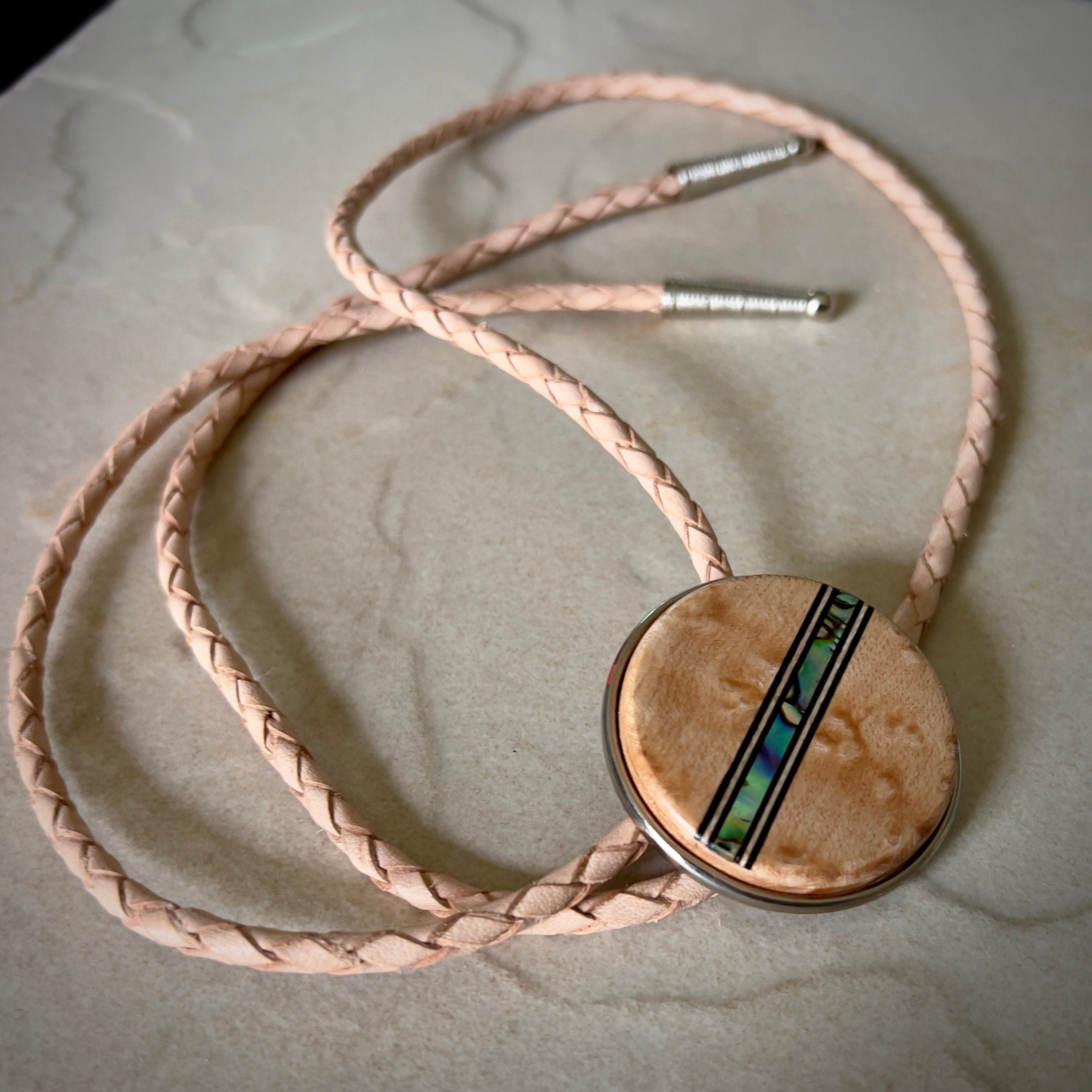 Bolo tie with a round wooden slide with iridescent vertical inlay strip and tan braided cords on a wooden surface.