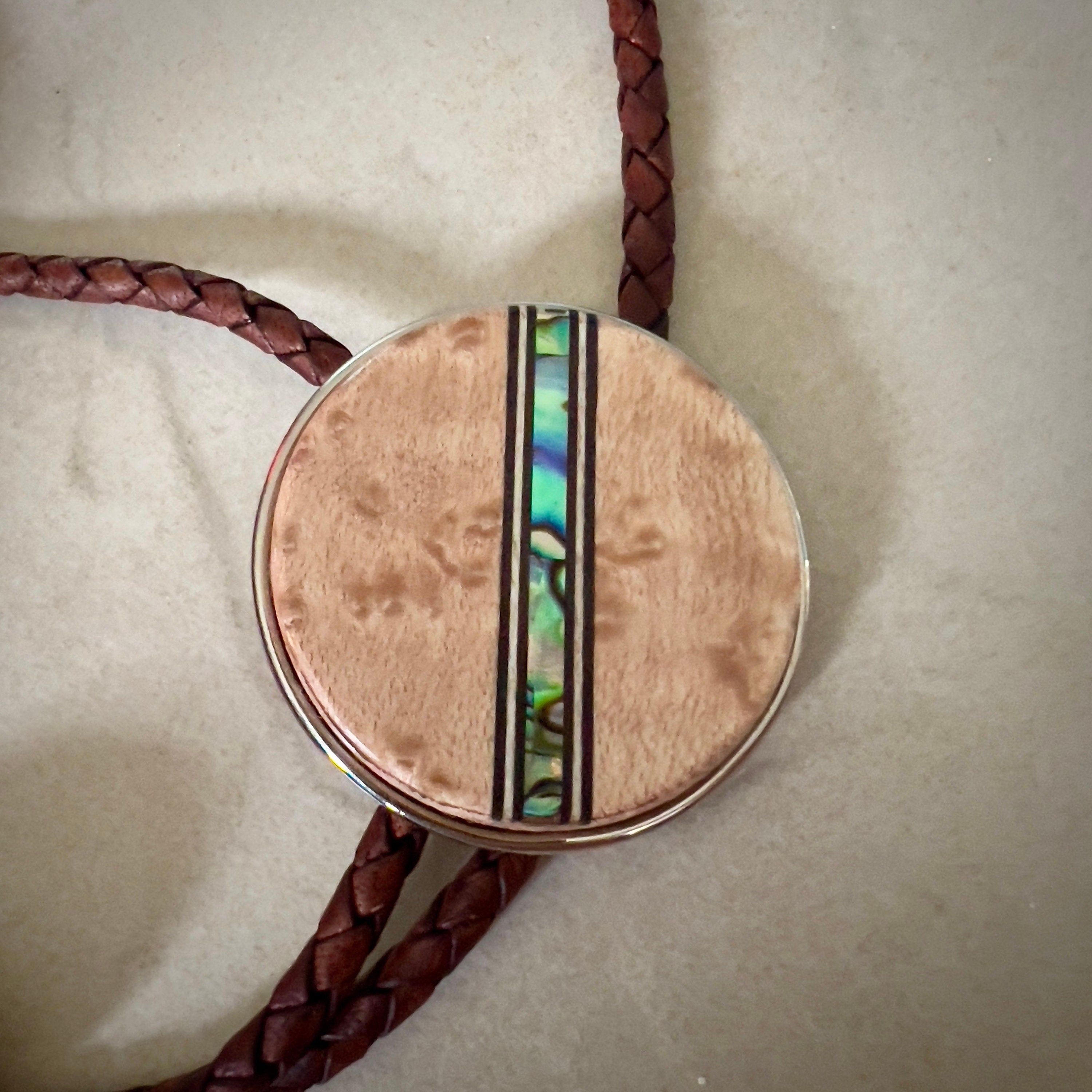 Bolo tie with a round wooden slide with iridescent vertical inlay strip and brown braided cords on a wooden surface.