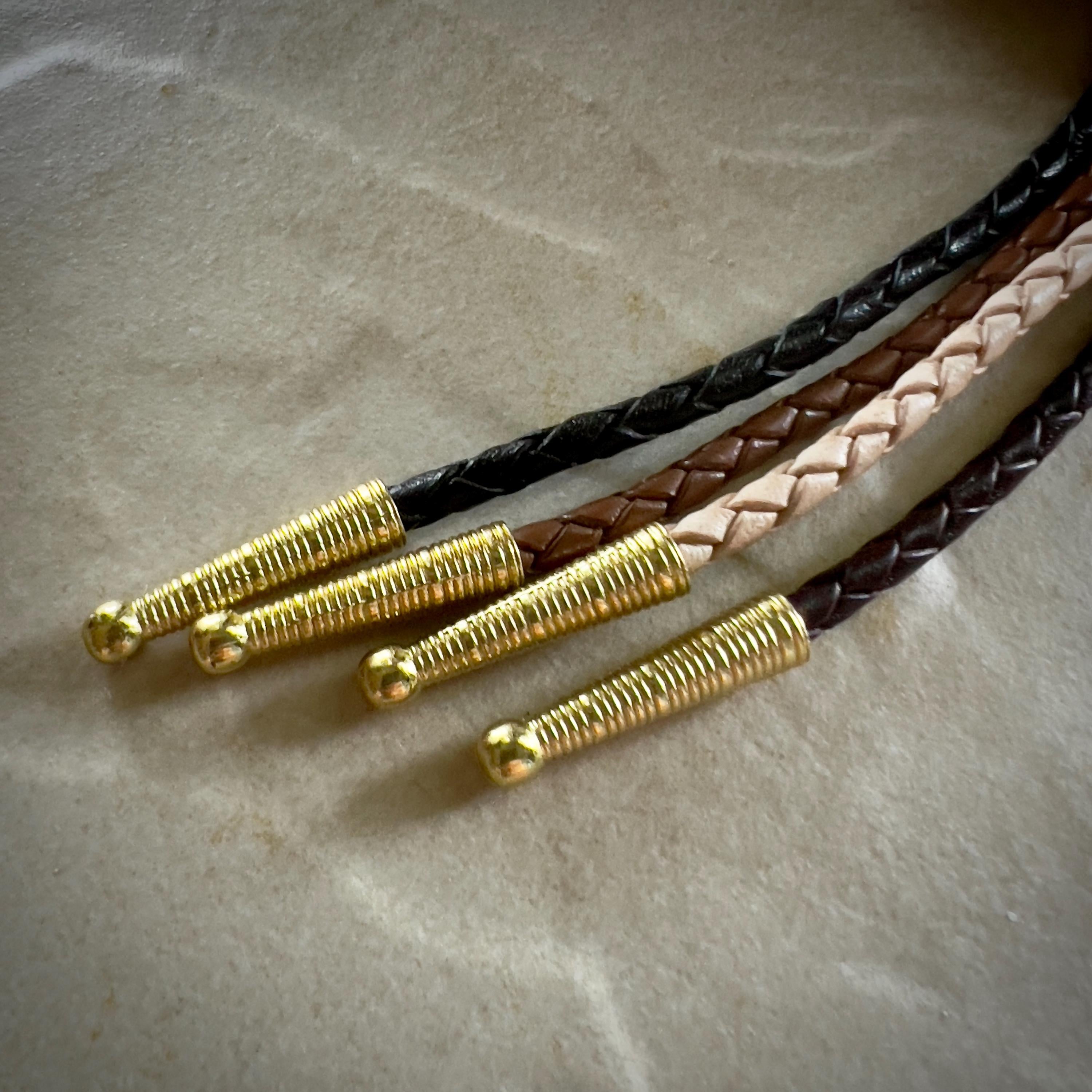 Four stripes of black, natural, saddle and brown braided leather rope with gold tips on a textured beige surface.