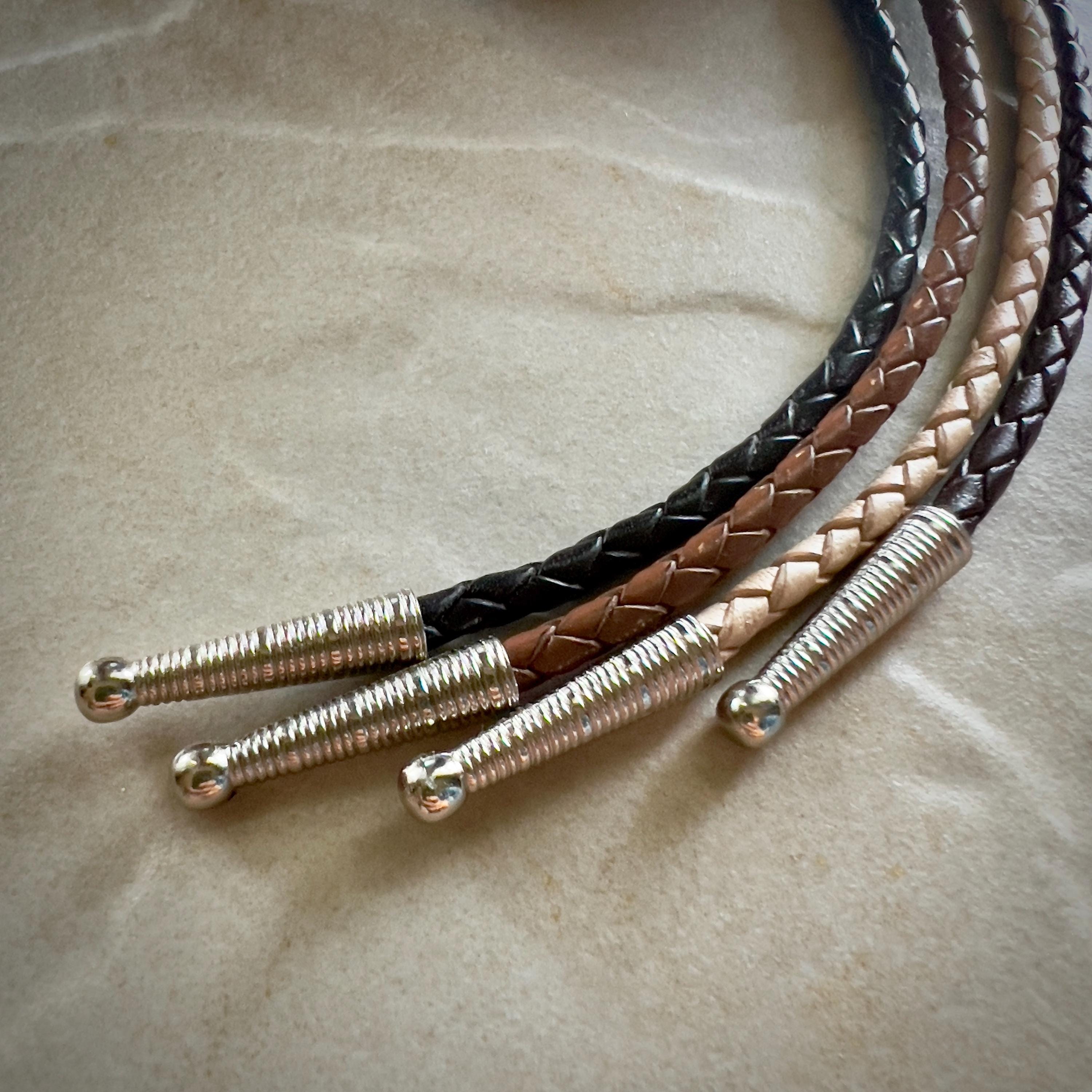 Four stripes of black, natural, saddle and brown braided leather rope with silver tips on a textured beige surface.