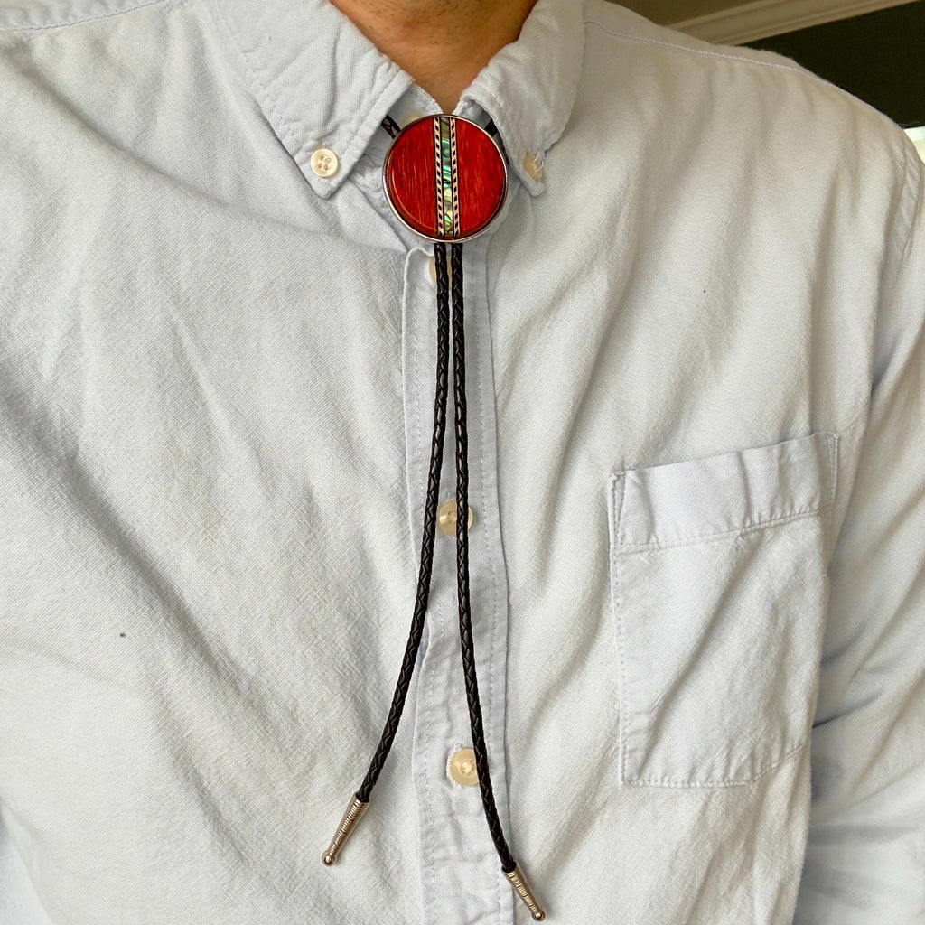 A person wearing a bolo tie with a red, circular slide featuring decorative shell inlay. The black braided leather cords of the tie are fastened with silver metal tips and hang over a light blue button-up shirt.