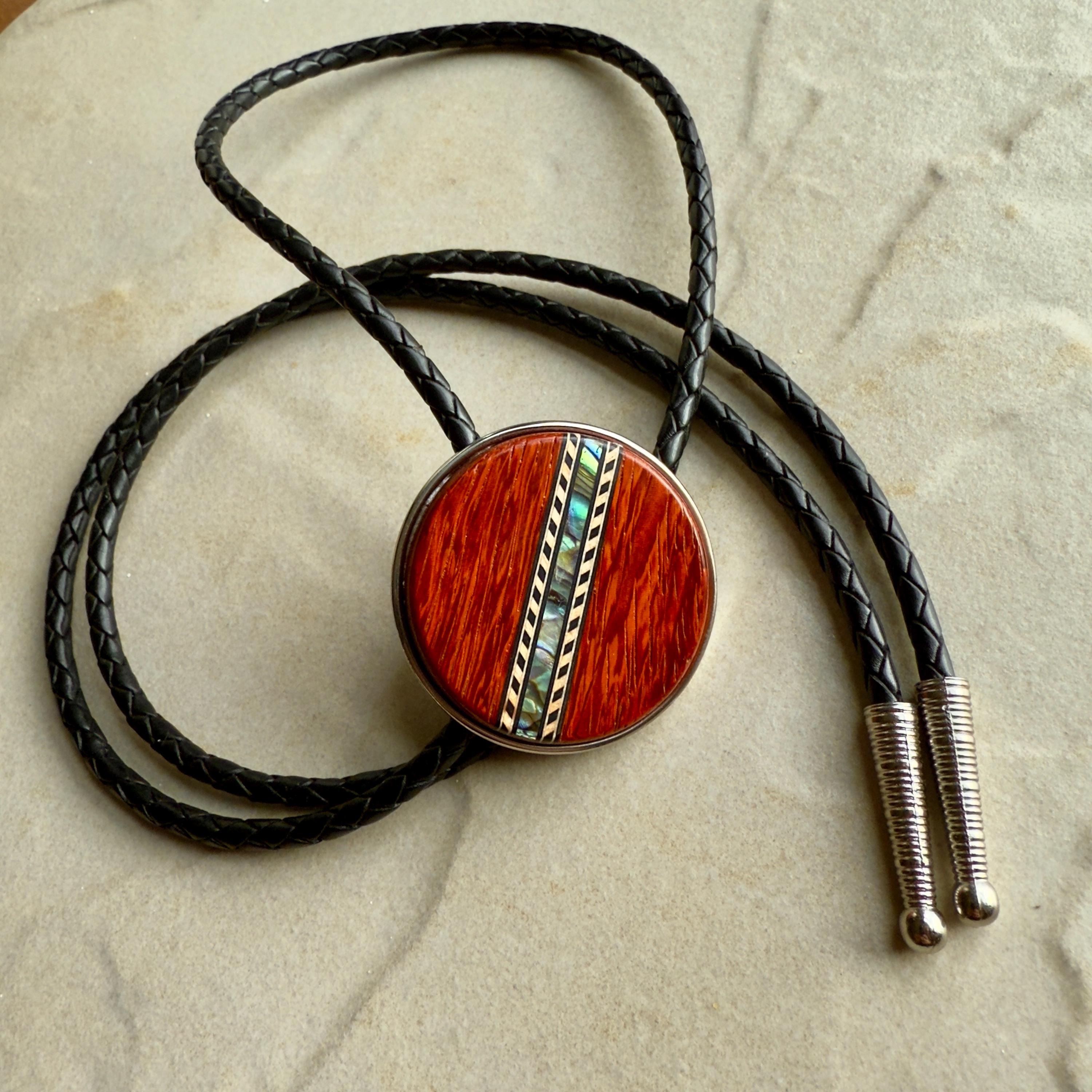 A black braided leather bolo tie with a round, red wood center with silver tips on a stone background.