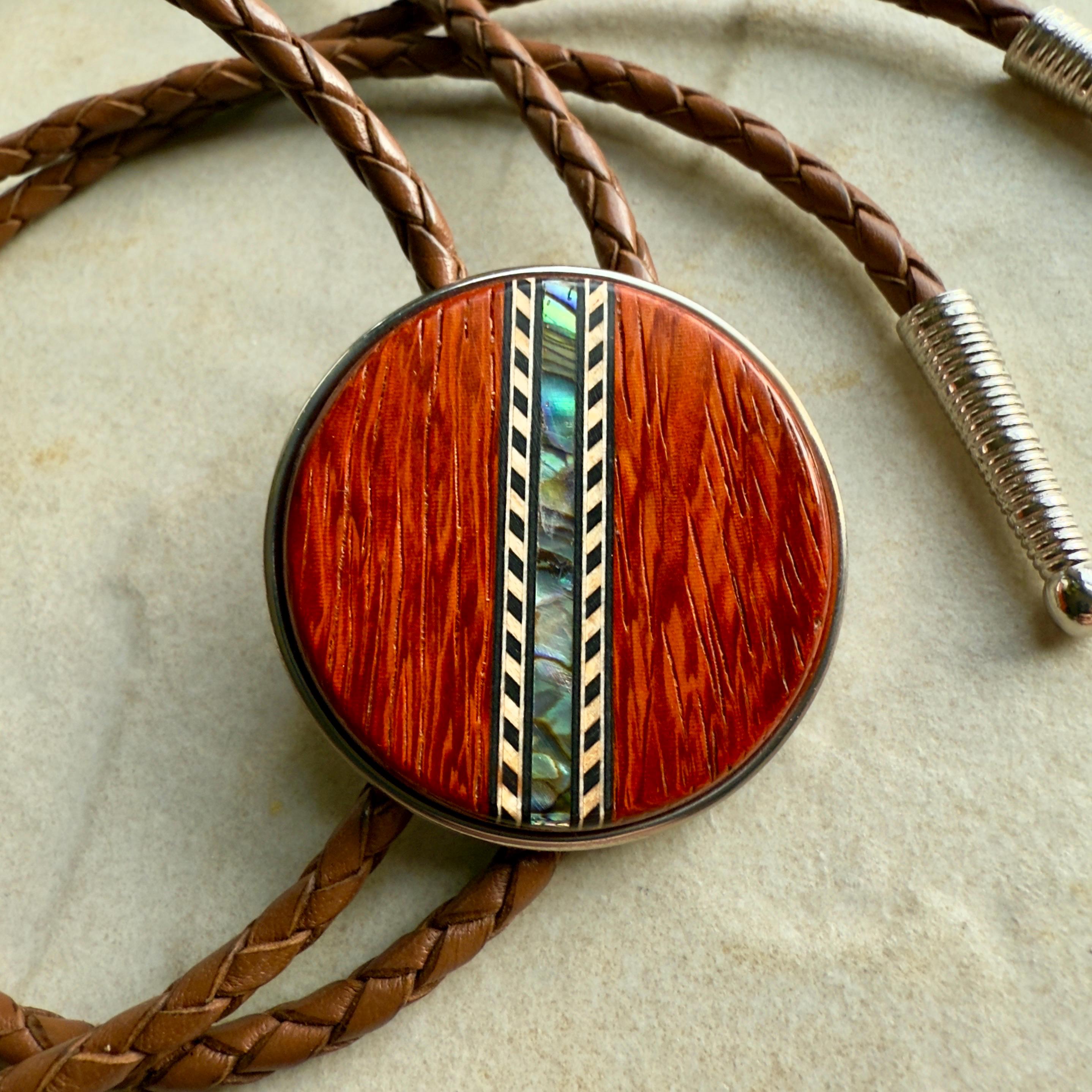 A round, wooden bolo side with a central inlay strip of shell inlay, attached to a brown braided cord with a silver metal tip.