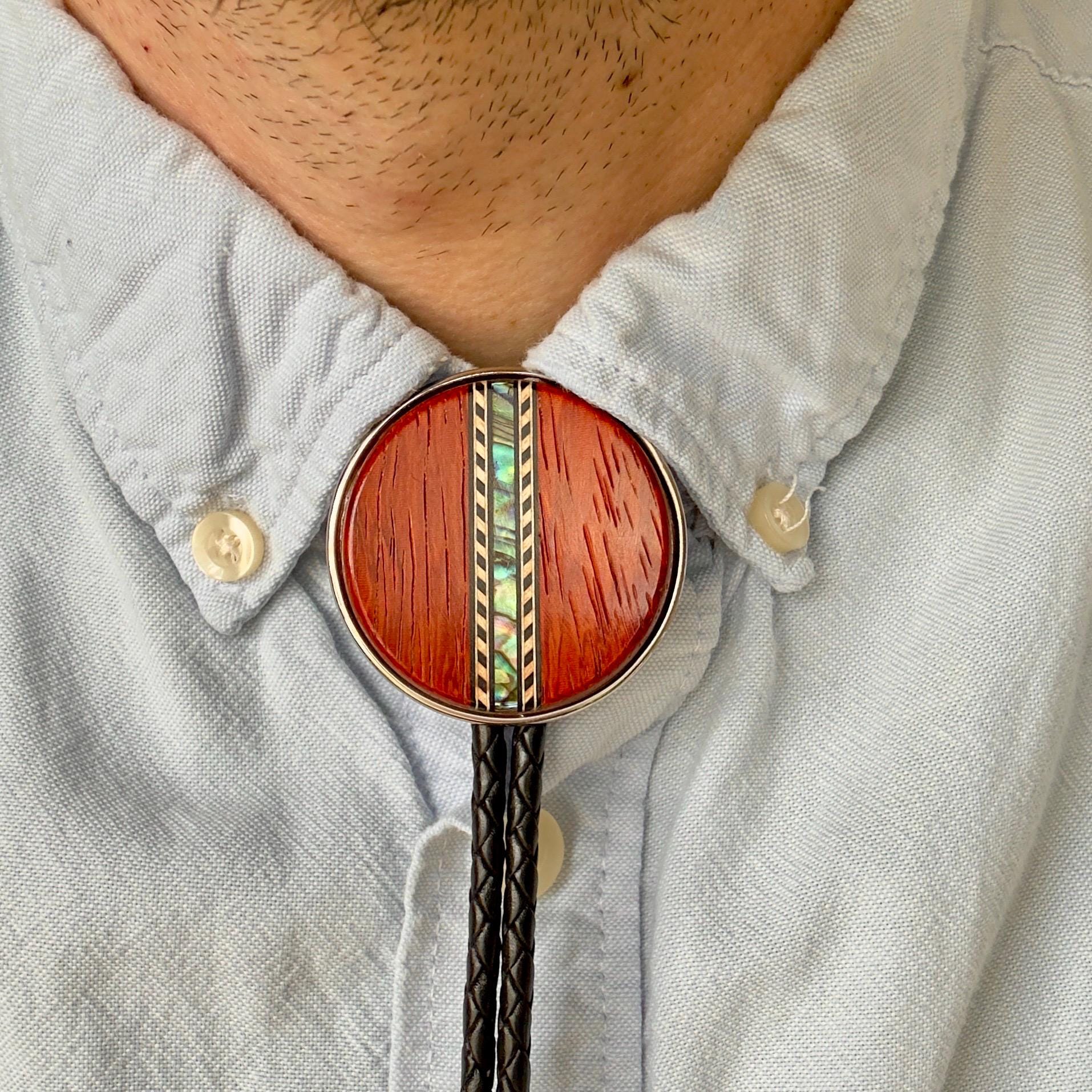 A person wearing a bolo tie with a red, circular slide featuring decorative shell inlay. The black braided leather cords of the tie are hung over a light blue button-up shirt.