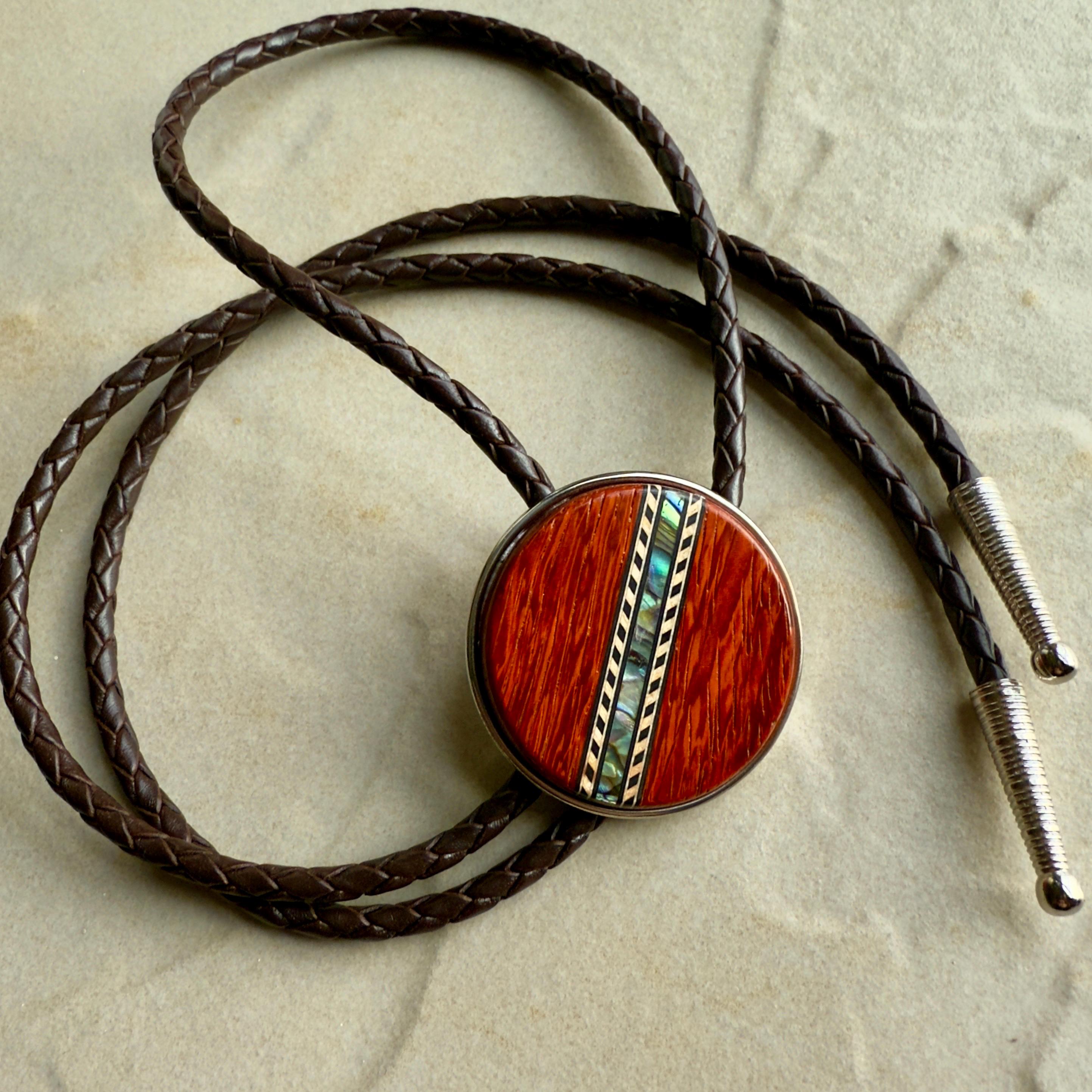 A brown leather braided bolo tie with a round, red wood center with silver tips on a stone background.