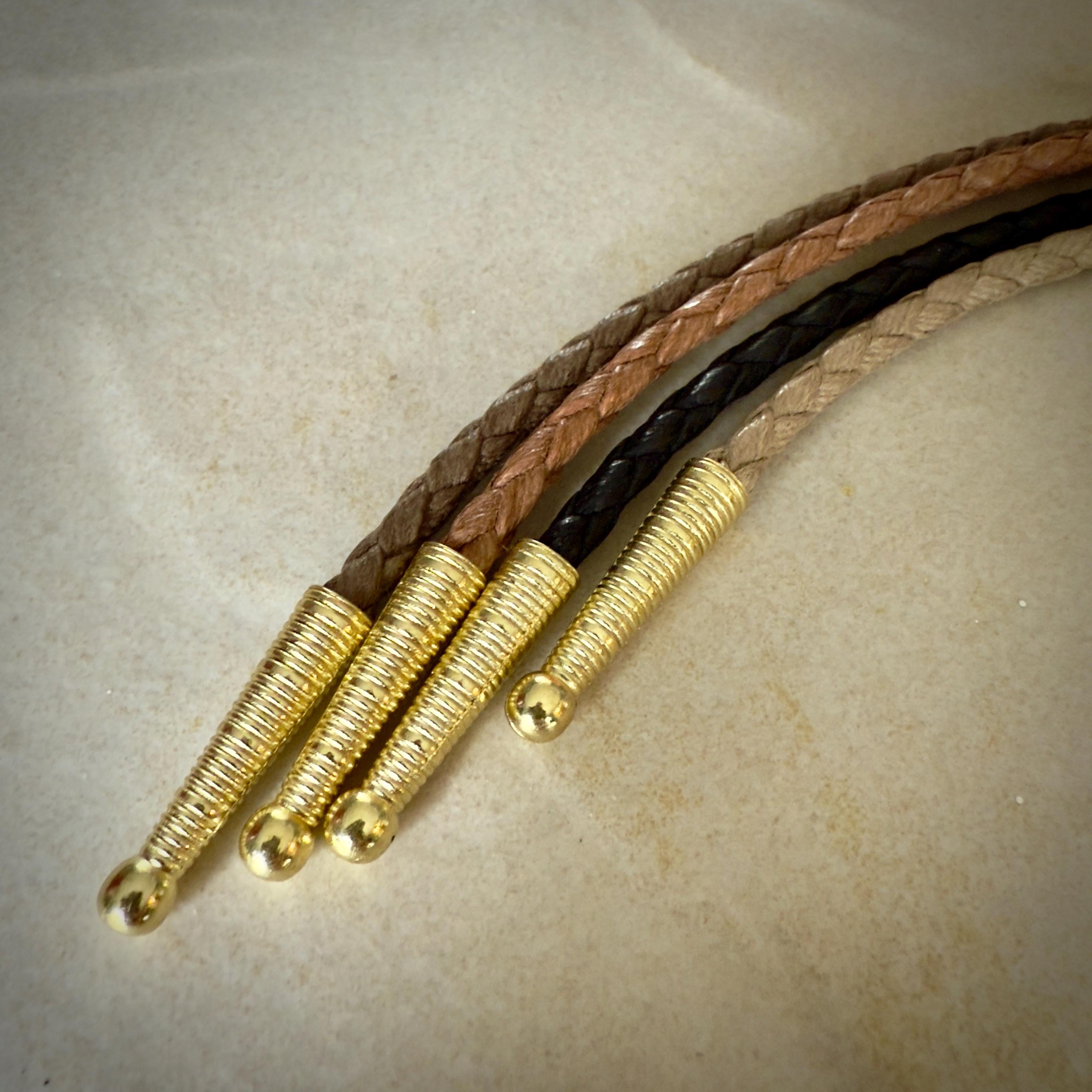 Four stripes of coffee, saddle, black and sand braided leather rope with gold tips on a textured beige surface.