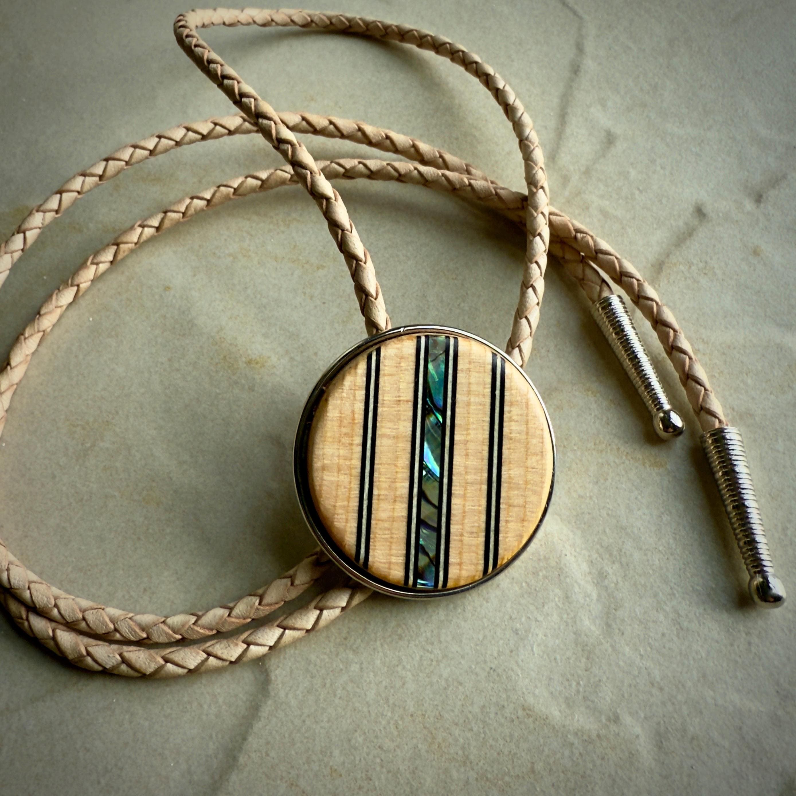A bolo tie featuring a round wooden slide with vertical inlays of black lines and iridescent abalone shell, adding a colorful accent. The braided natural leather cords end in sleek silver tips, displayed on a neutral stone surface for contrast.