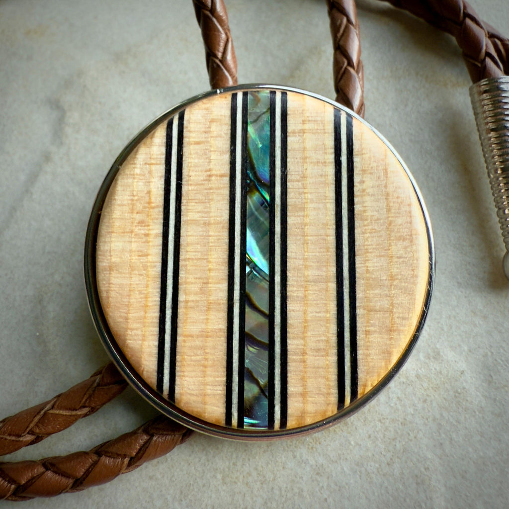 A close-up of a bolo tie's wooden slide featuring a natural wood finish, accented with vertical black lines and a shimmering abalone inlay at its center. The braided brown leather cords and silver tips are partially visible around the centerpiece.