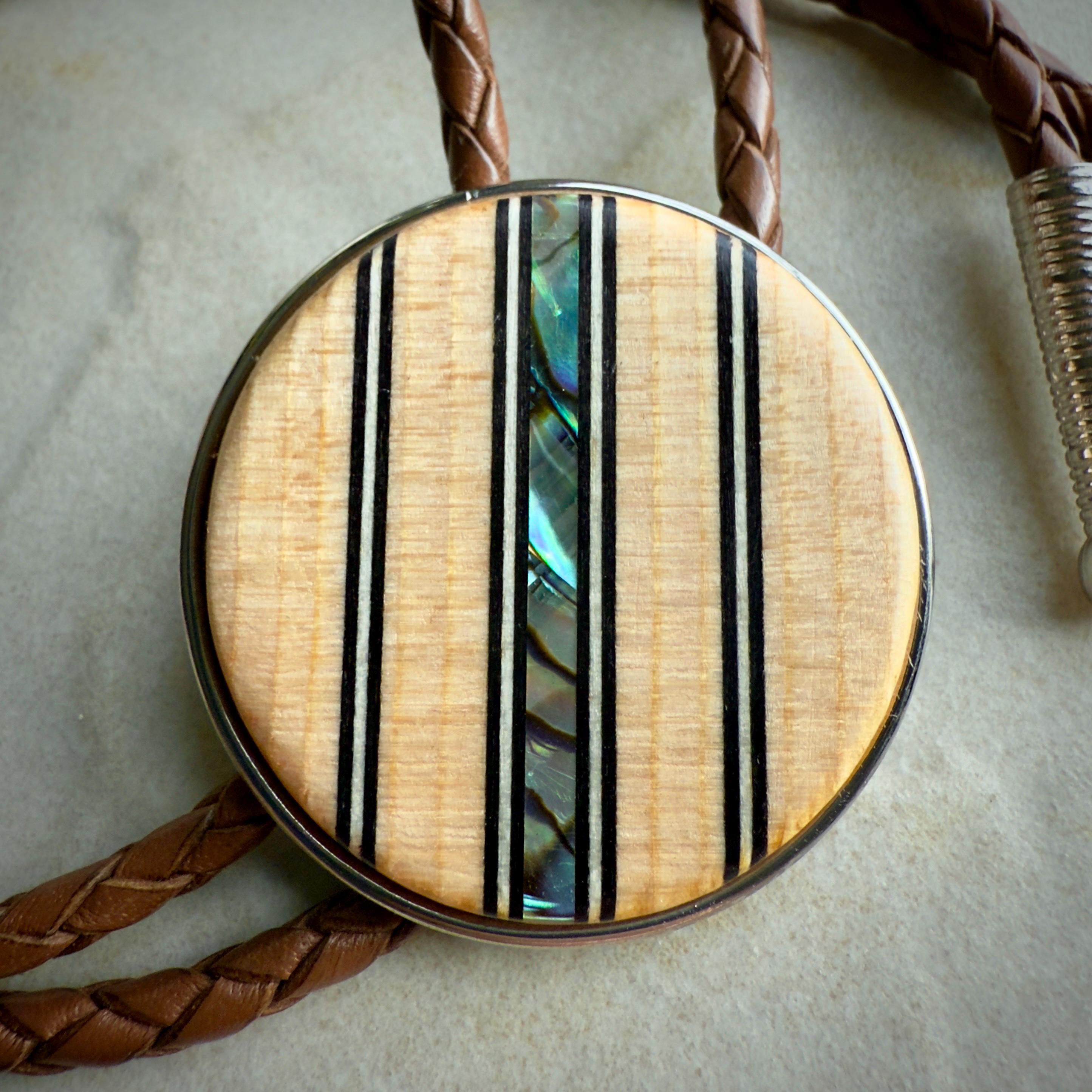 A close-up of a bolo tie's wooden slide featuring a natural wood finish, accented with vertical black lines and a shimmering abalone inlay at its center. The braided brown leather cords and silver tips are partially visible around the centerpiece.