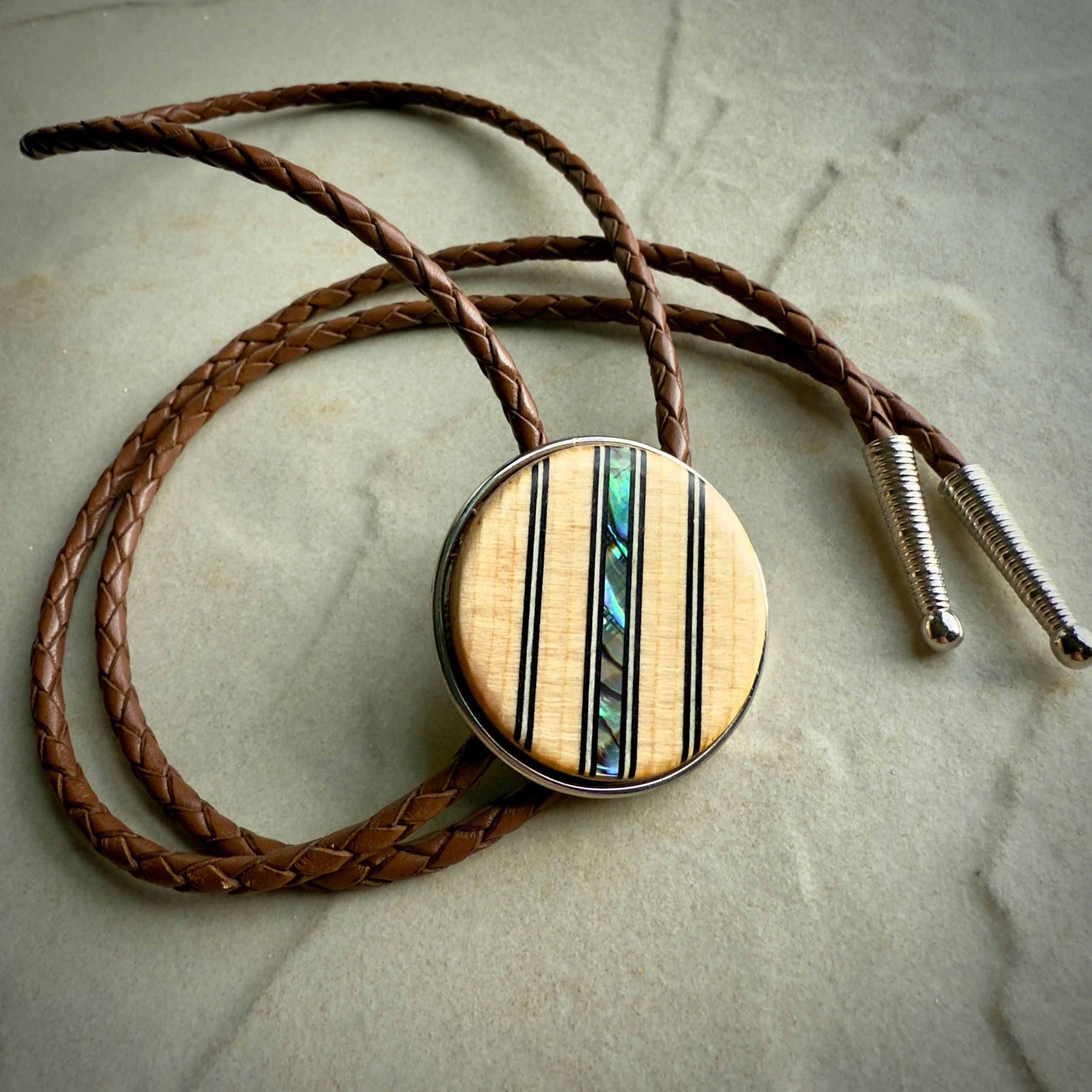A bolo tie featuring a round wooden slide with vertical inlays of black lines and iridescent abalone shell, adding a colorful accent. The braided saddle leather cords end in sleek silver tips, displayed on a neutral stone surface for contrast.
