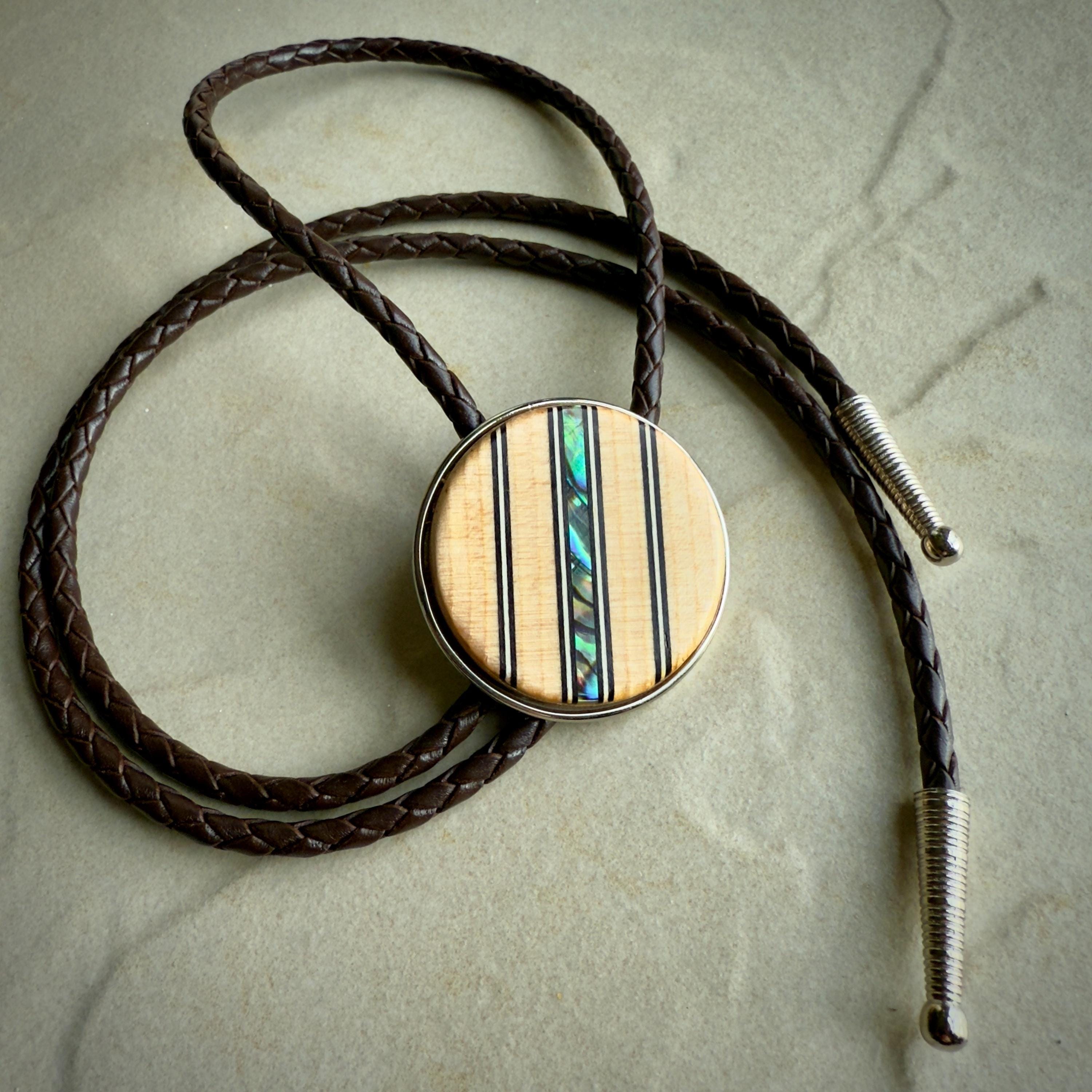 A bolo tie featuring a round wooden slide with vertical inlays of black lines and iridescent abalone shell, adding a colorful accent. The braided dark brown leather cords end in sleek silver tips, displayed on a neutral stone surface for contrast.