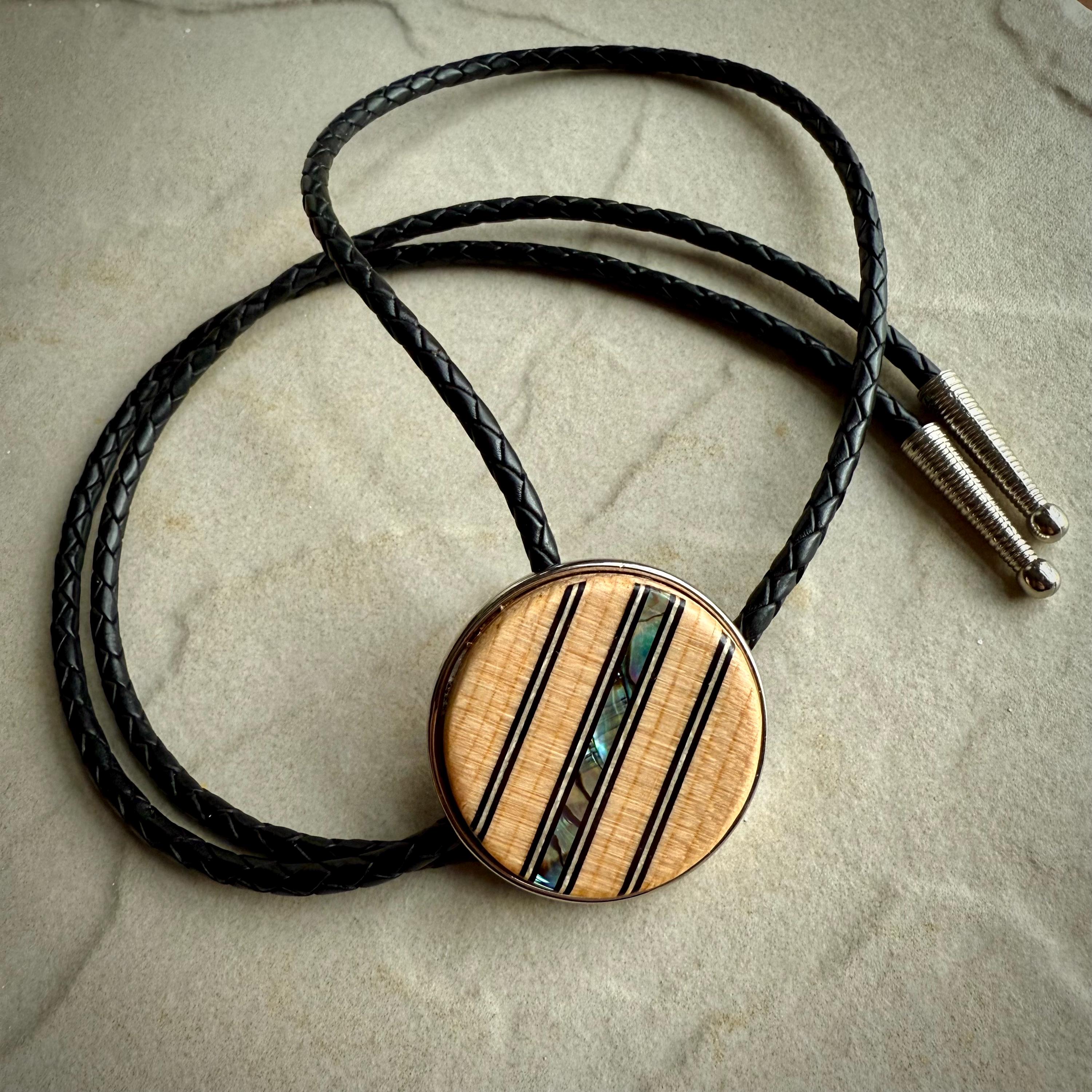 A bolo tie featuring a round wooden slide with vertical inlays of black lines and iridescent abalone shell, adding a colorful accent. The braided black leather cords end in sleek silver tips, displayed on a neutral stone surface for contrast.