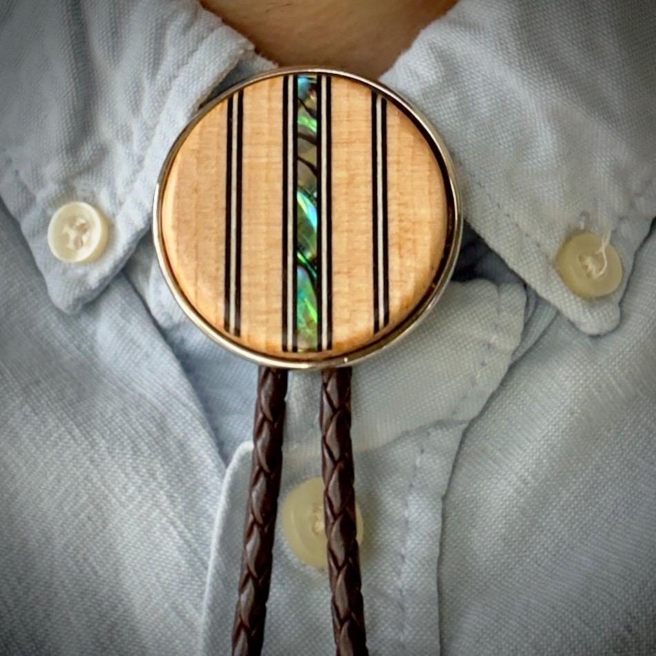 A close-up of a bolo tie worn with a light blue button-down shirt. The wooden slide features vertical black lines and a shimmering abalone inlay. The dark brown braided leather cords hang neatly below.