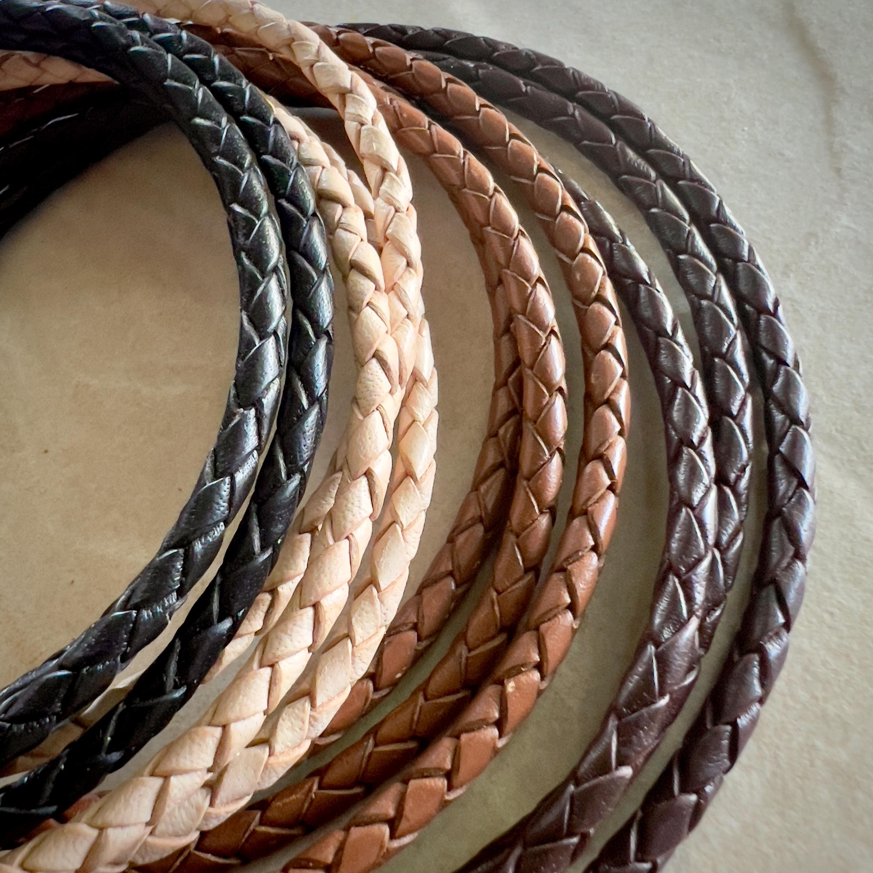 Close-up of several coiled braided leather cords arranged in a semi-circle on a stone surface. The cords are in varying shades, including black, natural, saddle, and dark brown, showcasing their texture and craftsmanship.